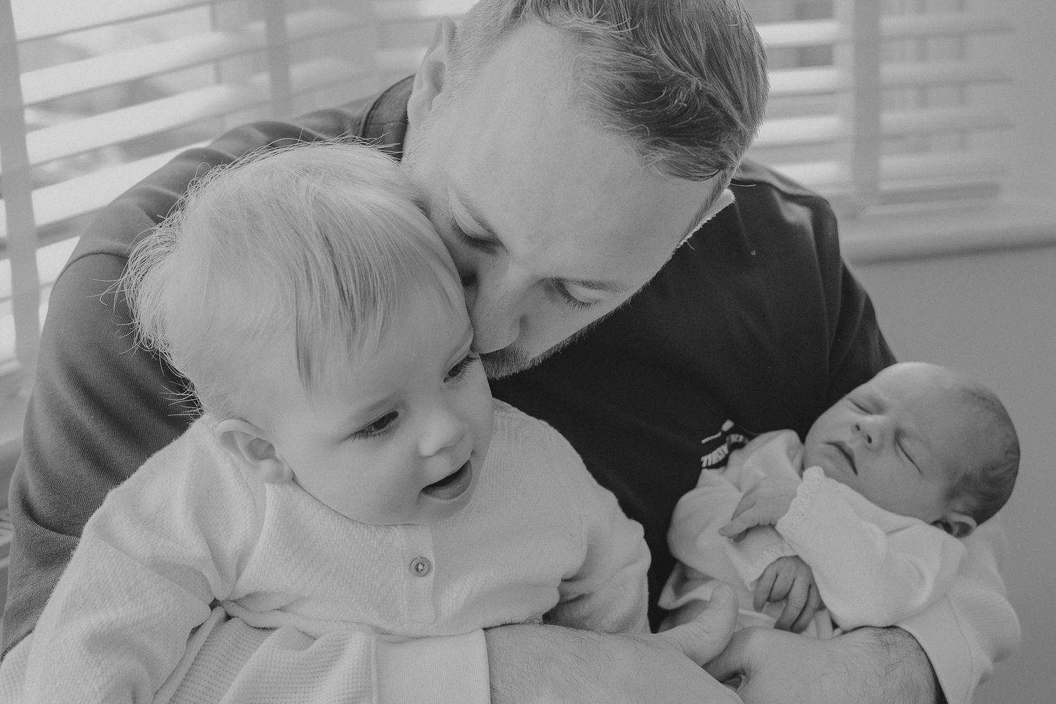Black and white photo of a man kissing a toddler while holding a newborn baby.