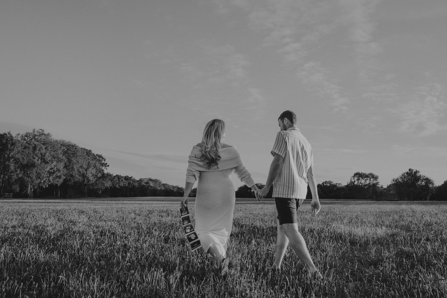 Black and white photo of a couple holding hands walking through a grassy field with trees in the background.