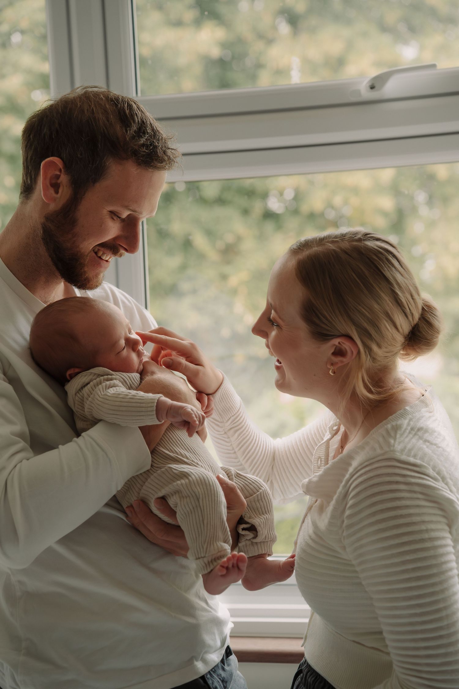 A series of tender moments between parents and their newborn baby by a window in natural light.