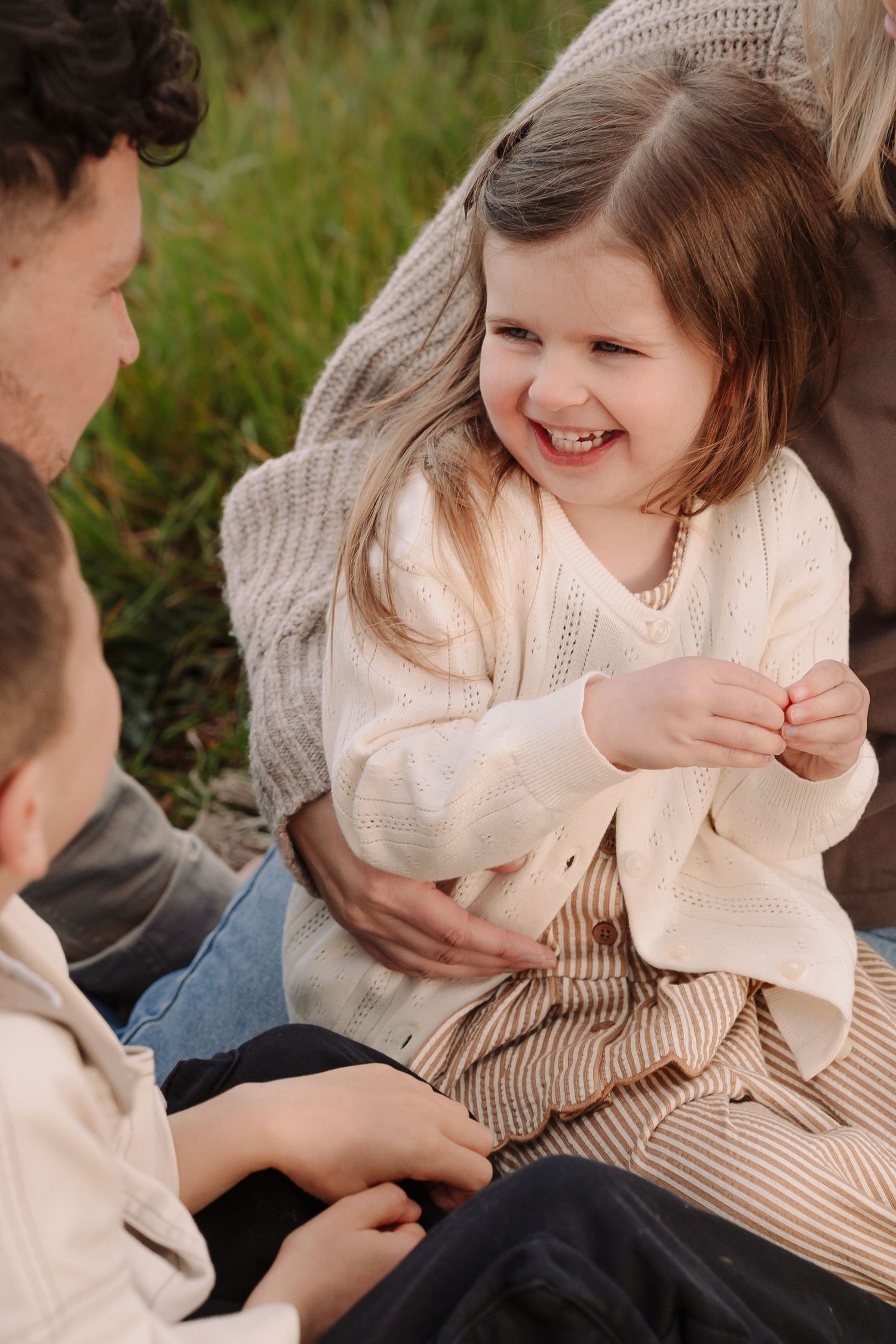 A laughing little girl in a cream knit cardigan sits outdoors with family during a warm candid moment.