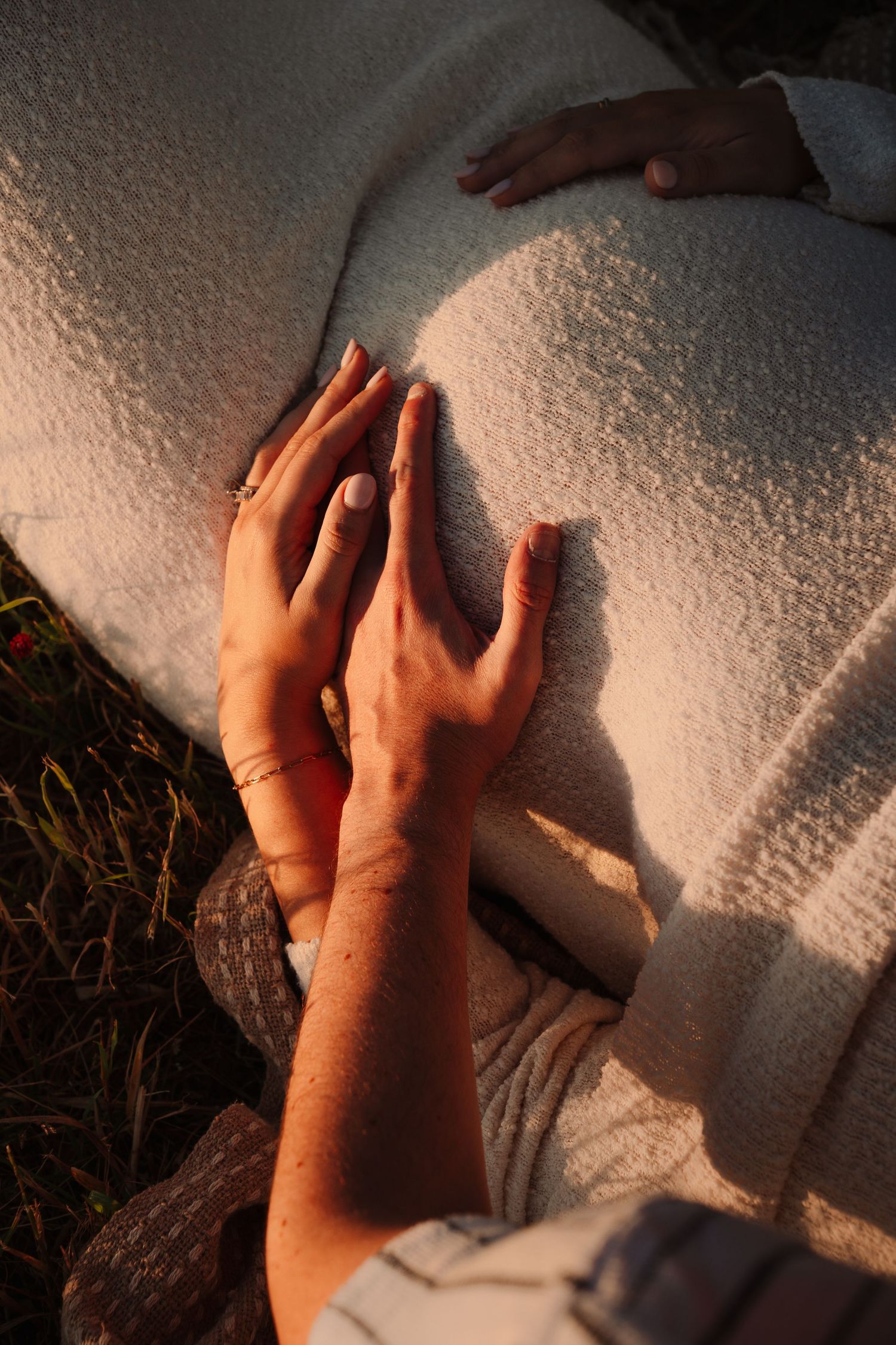 Hands gently resting on a pregnant belly wrapped in white fabric, bathed in warm golden sunset light outdoors.