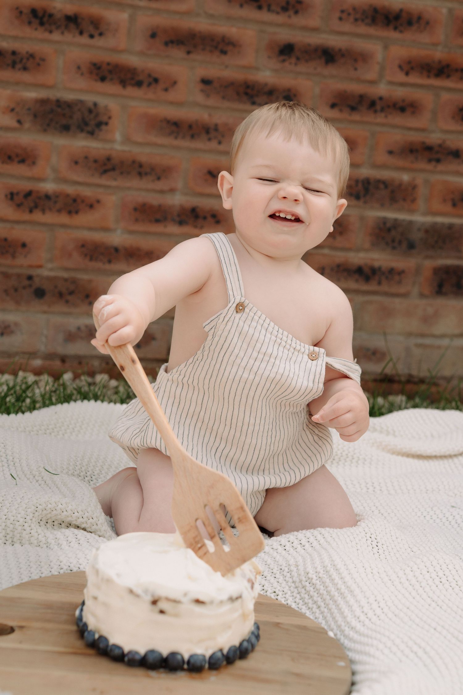 A baby in striped overalls sits on a white blanket playing with a wooden toy drum on a brick background.