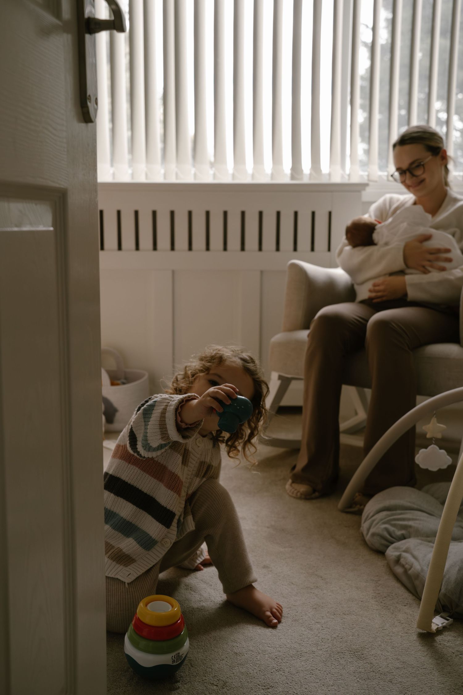 A toddler plays with a colorful stacking toy on the carpet while someone sits in an armchair in a cozy nursery.