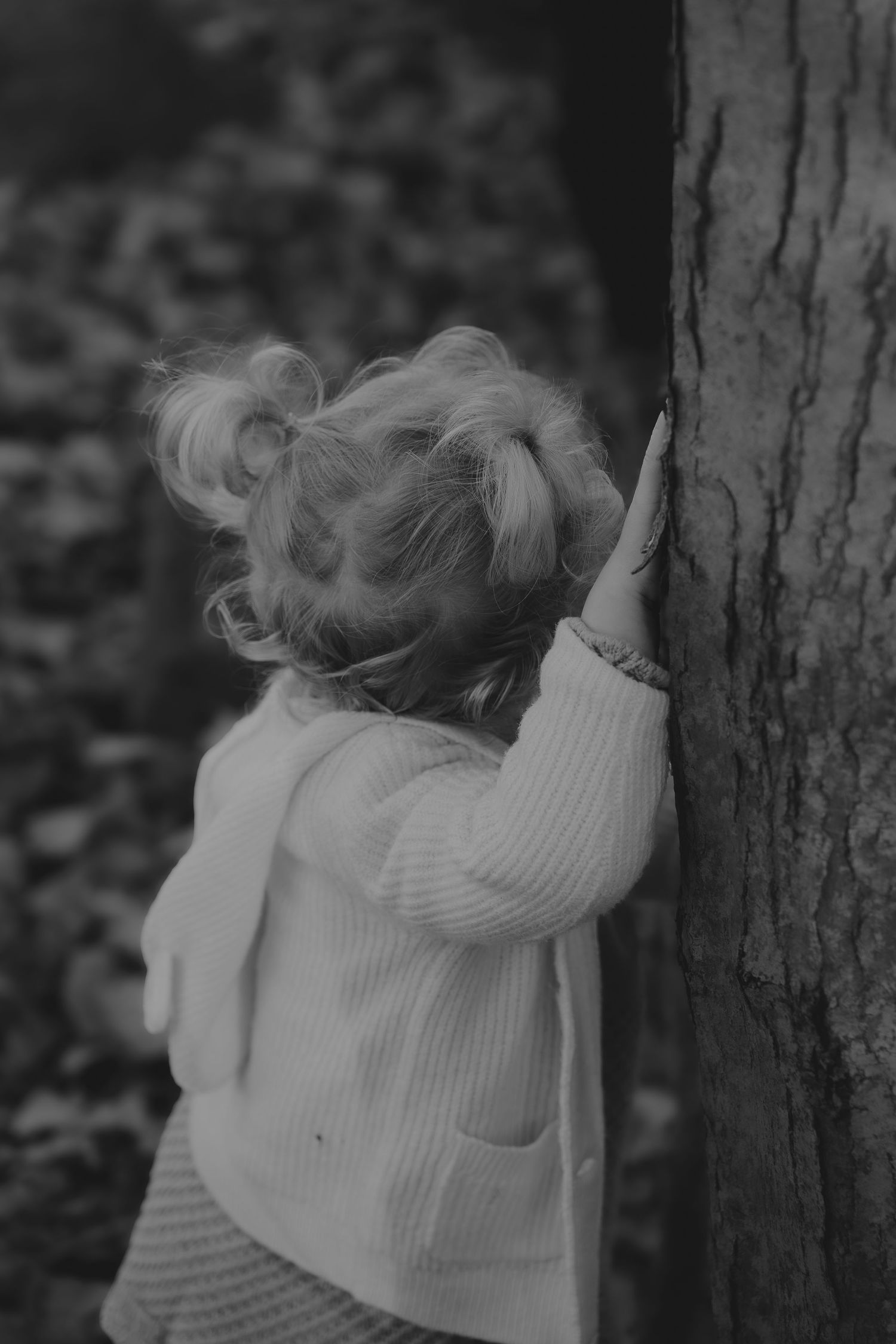 Child in white sweater reaches up to touch tree bark in black and white photo.