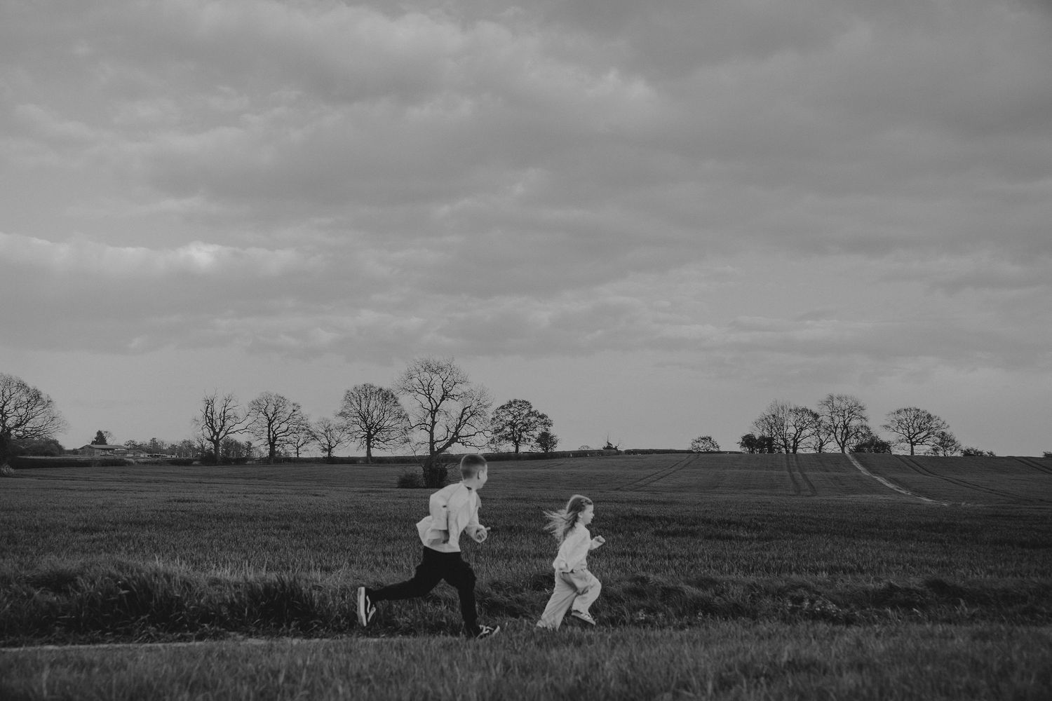 Black and white photo of a couple running through an open field with bare trees and cloudy skies in the background.