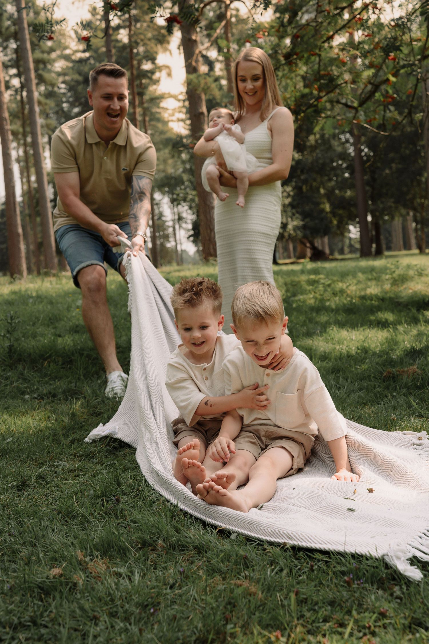Family enjoying playful moments together on a white blanket in a sunny park setting.