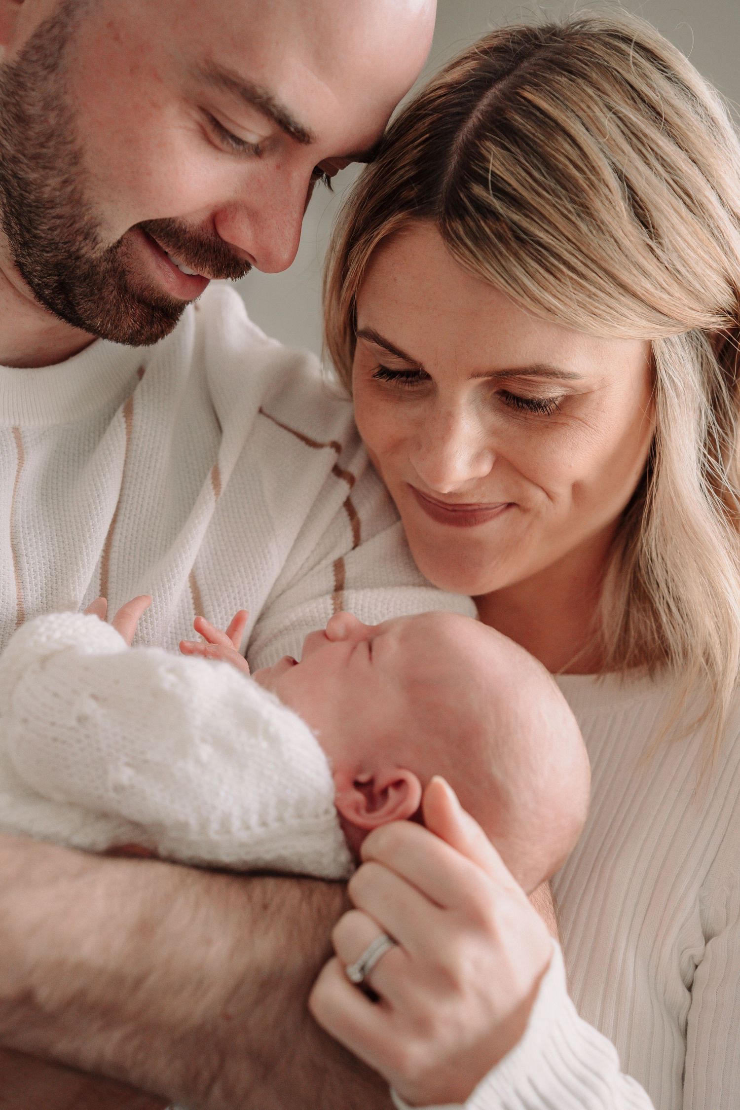 A tender family moment as parents in cream-colored sweaters gaze lovingly at their newborn baby wrapped in white.