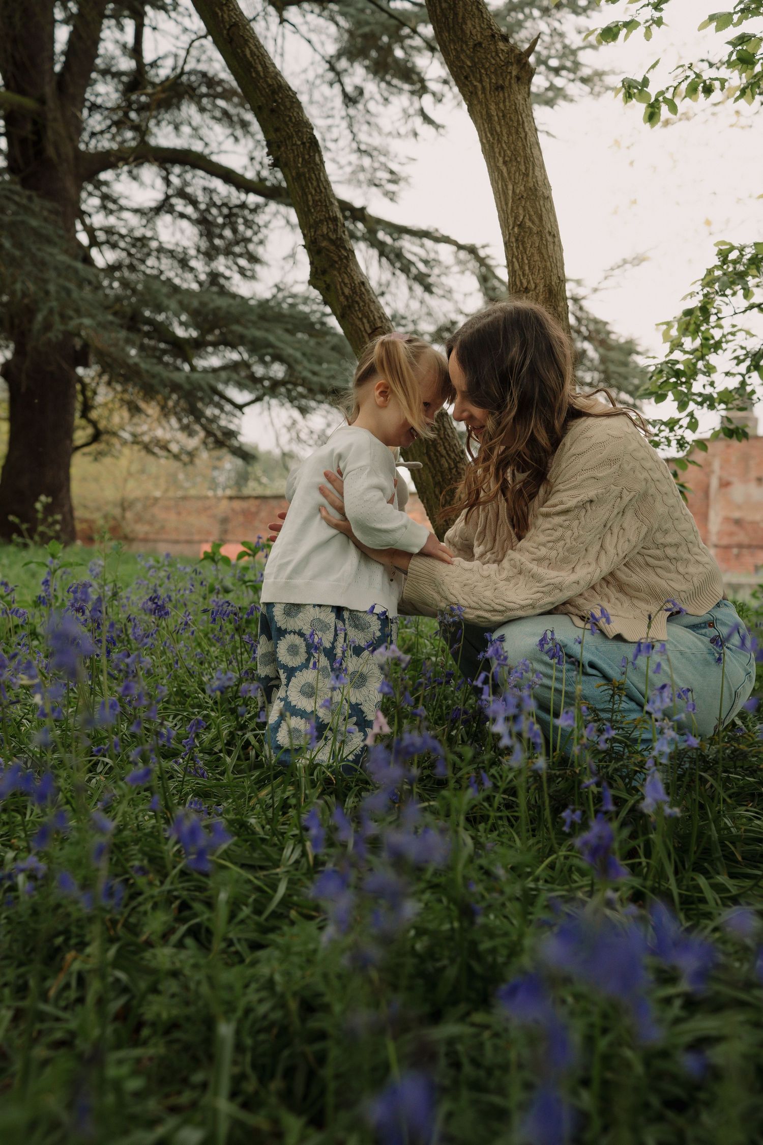 A couple sits together in a blooming field of purple wildflowers beneath tall trees during golden hour.