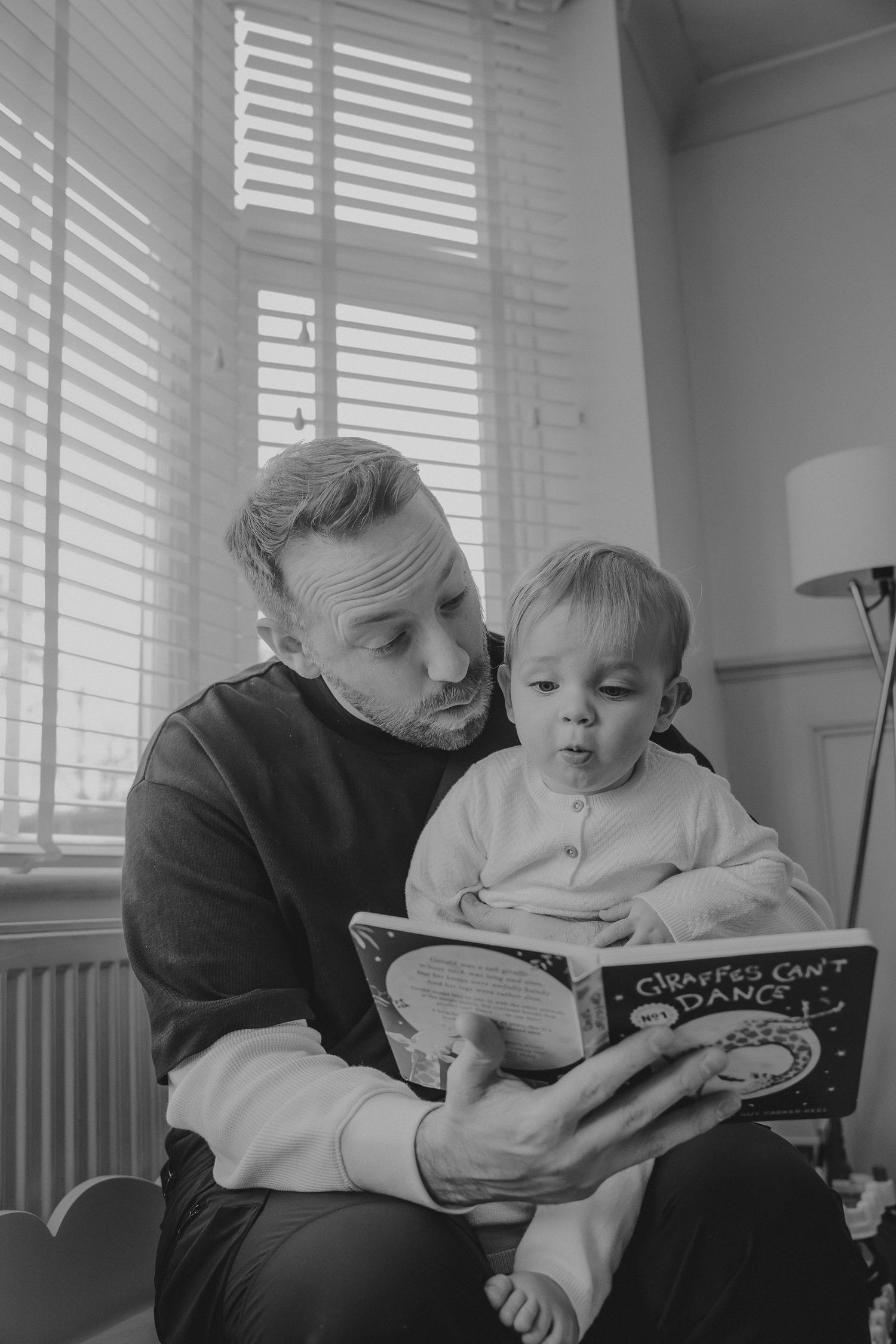 Father reads Giraffes Can't Dance book to toddler sitting on lap by window in black and white photo.