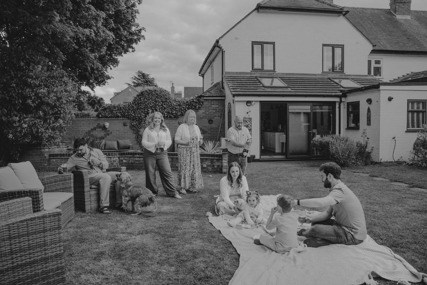 Black and white photo of a family gathering in a garden outside a semi-detached house on a cloudy day.