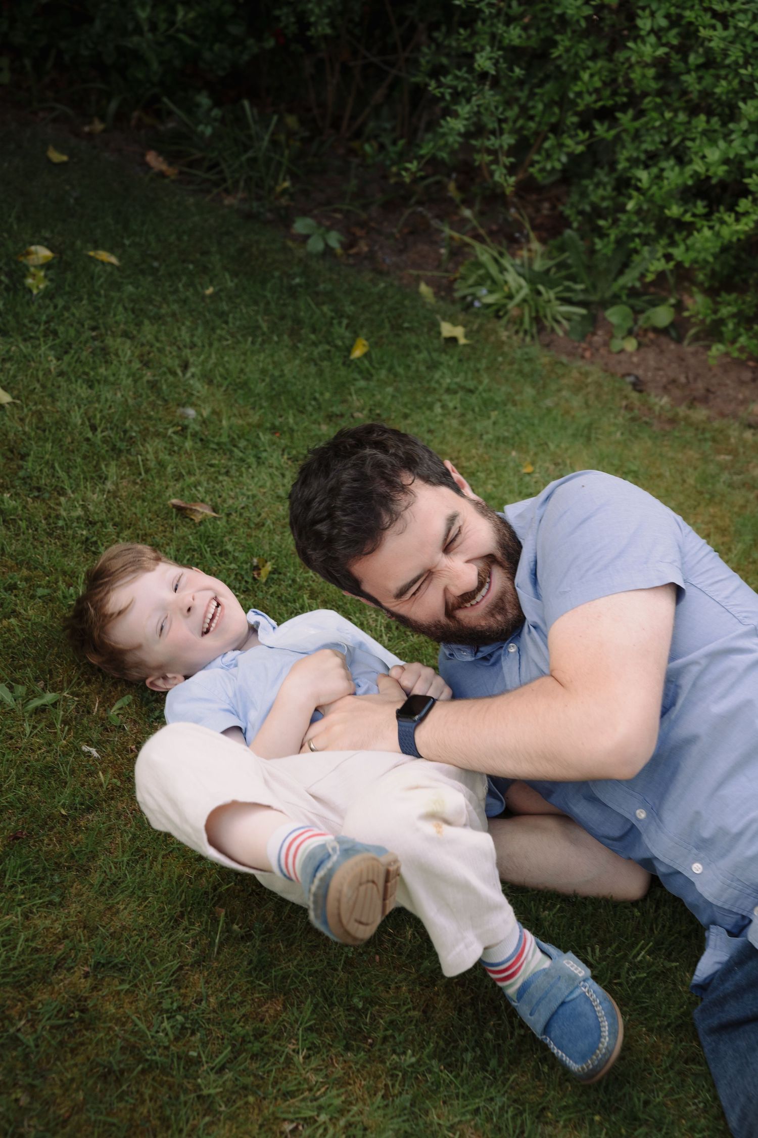 A laughing father plays with his happy baby on green grass in a garden, both wearing casual blue outfits.