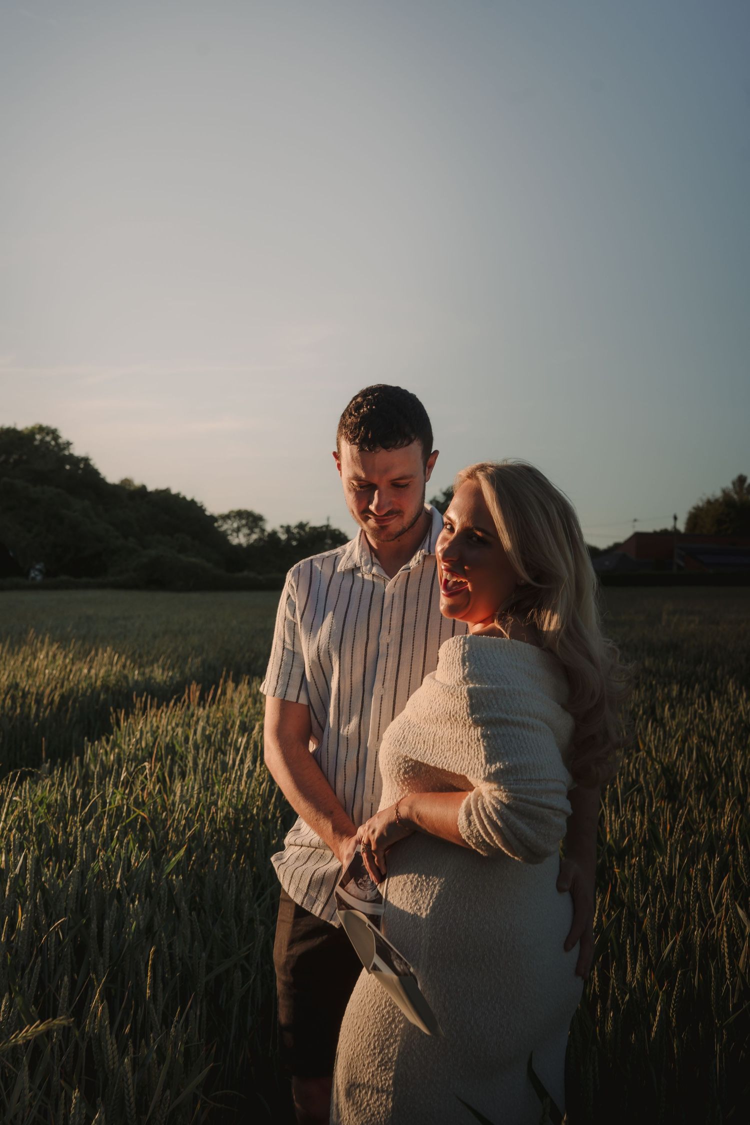 Couple standing in a golden field at sunset, woman in white dress smiling, man in striped shirt looking down.