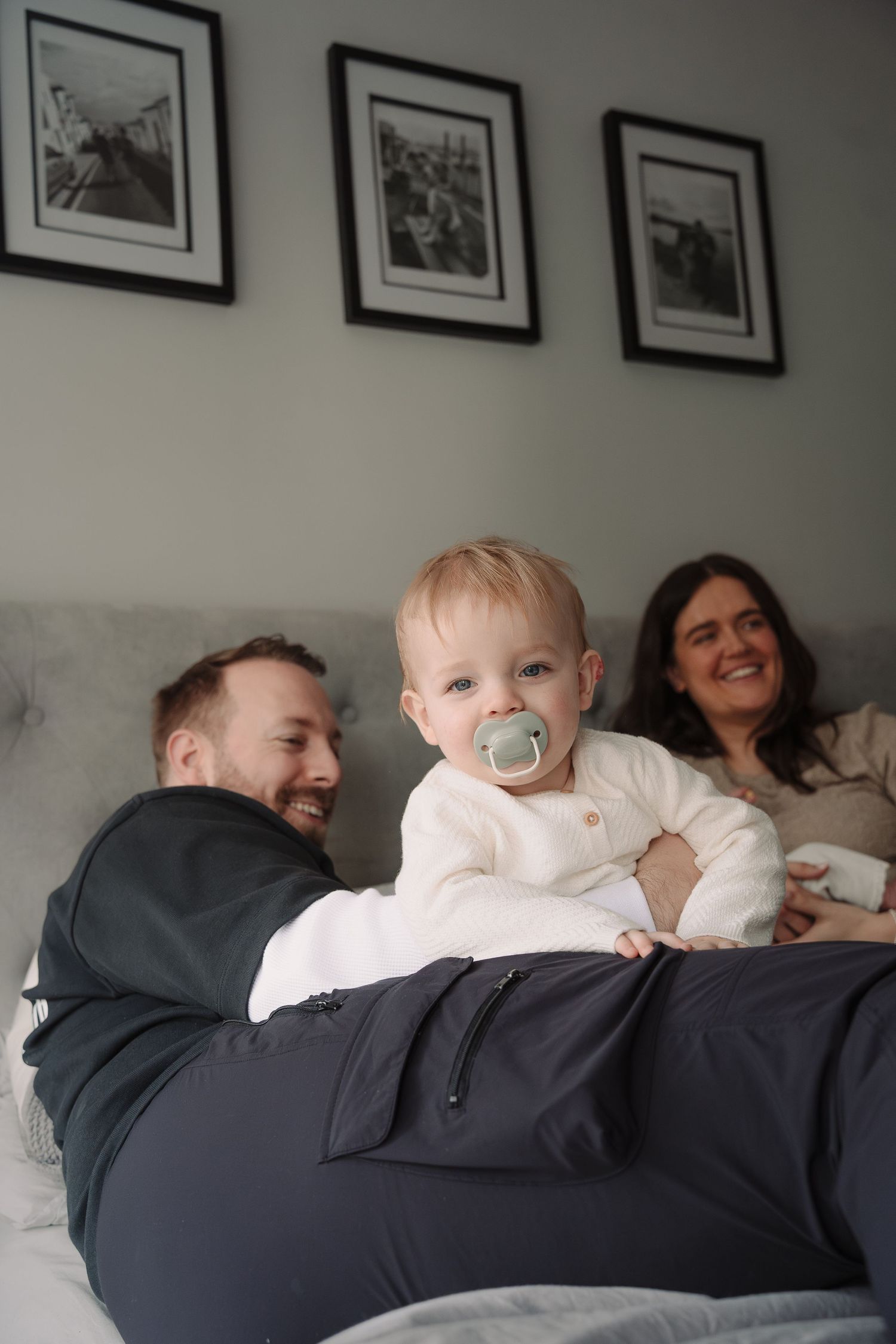 Happy family with smiling parents and baby with pacifier relaxing together on a bed.