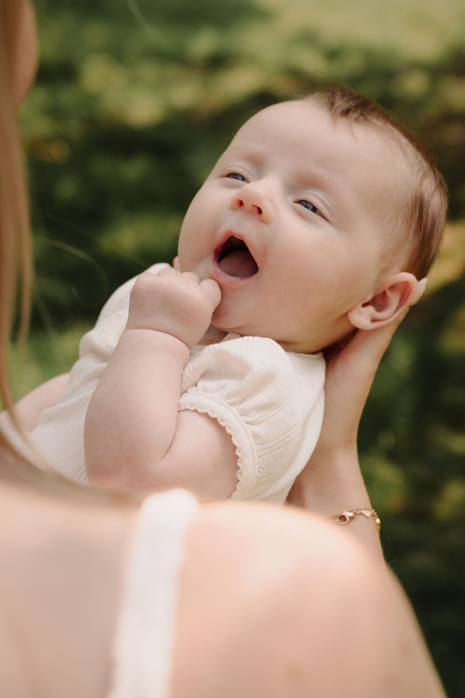 Baby in white dress yawns while being held outdoors with green foliage in background.
