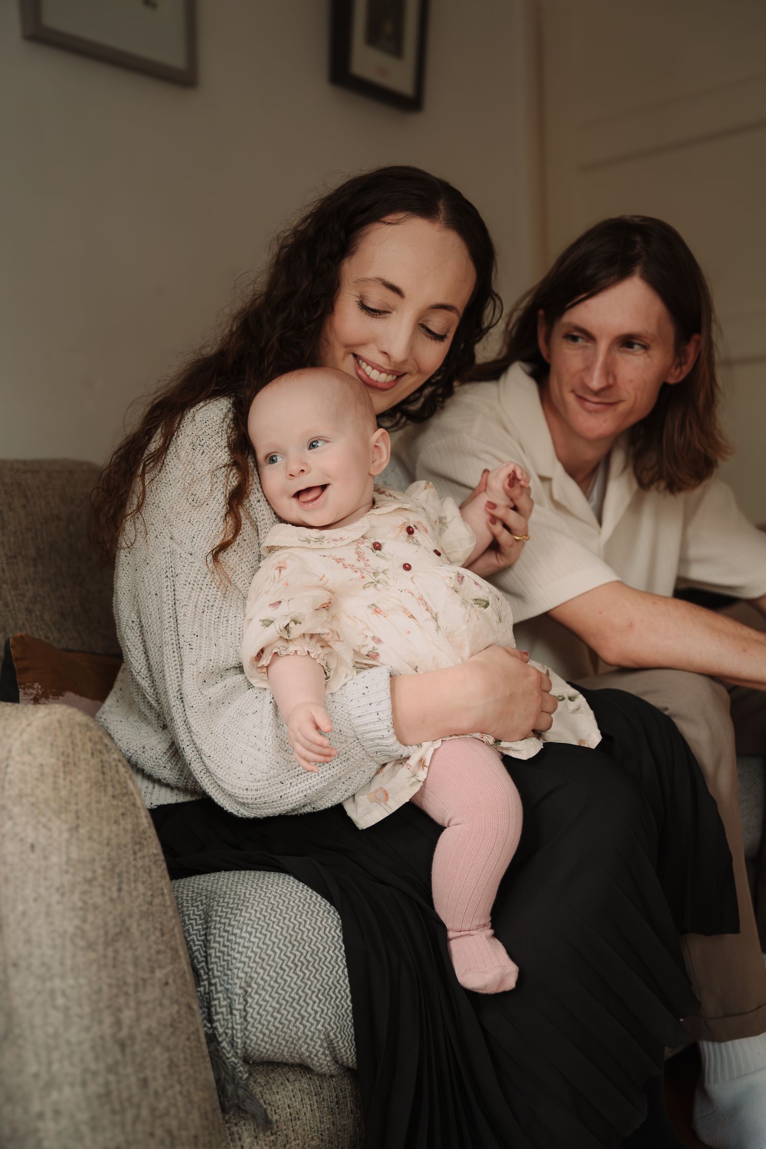 A happy family moment on the couch as parents share tender moments with their smiling baby in a floral dress.