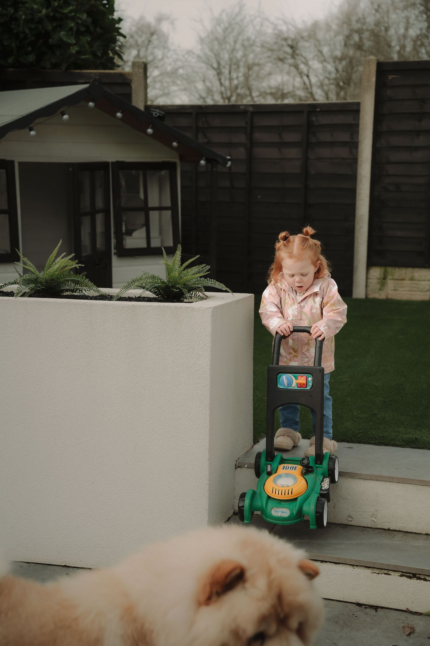 A child plays with a toy lawn mower in a modern backyard with white planters and dark fencing.