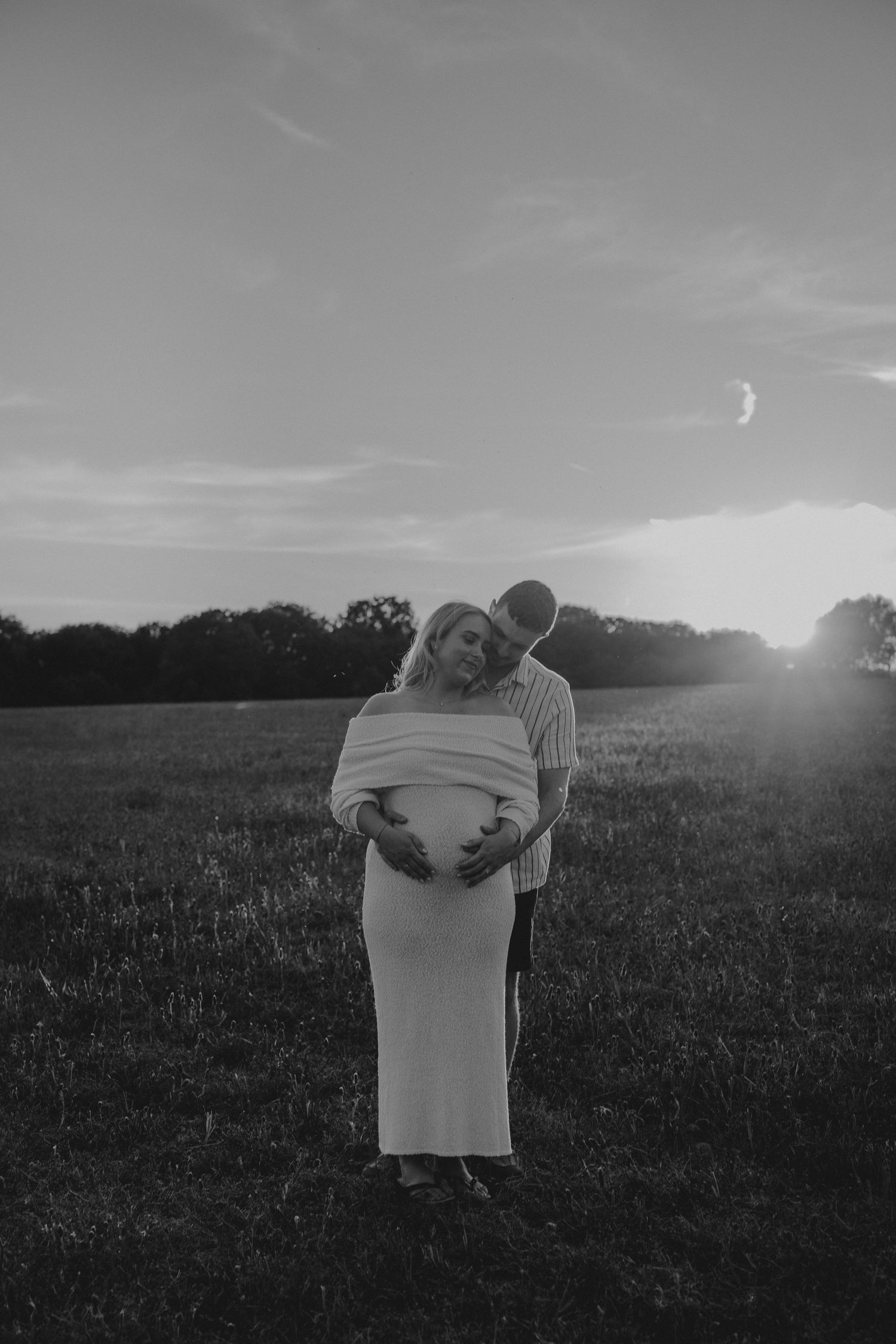 Black and white maternity photo of couple embracing in field at sunset, woman in elegant dress cradling baby bump.