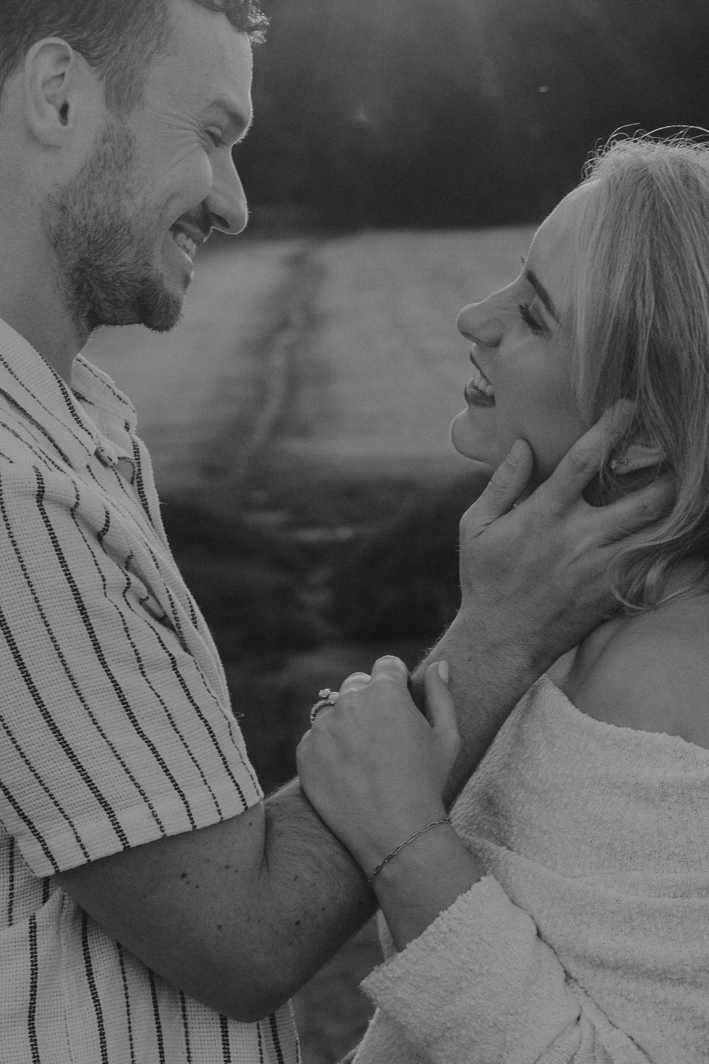 Black and white photo of a smiling couple sharing a tender moment, man gently holding womans face outdoors.