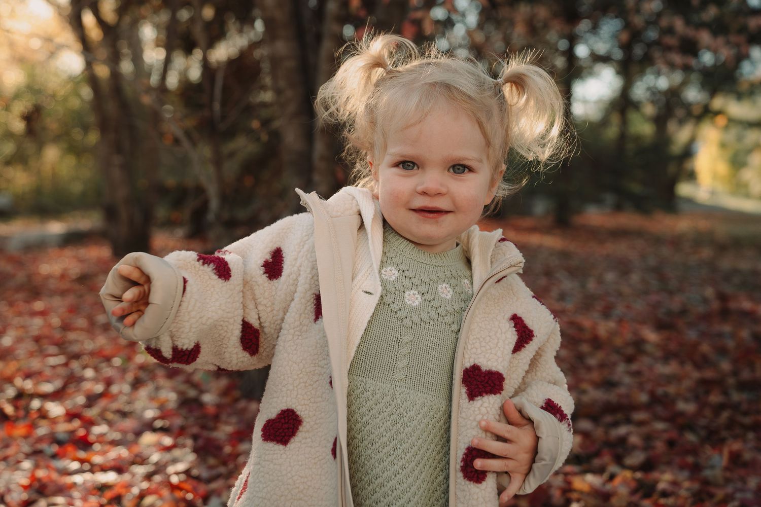 A child wearing a mint sweater and cream cardigan with burgundy hearts plays in fallen autumn leaves.