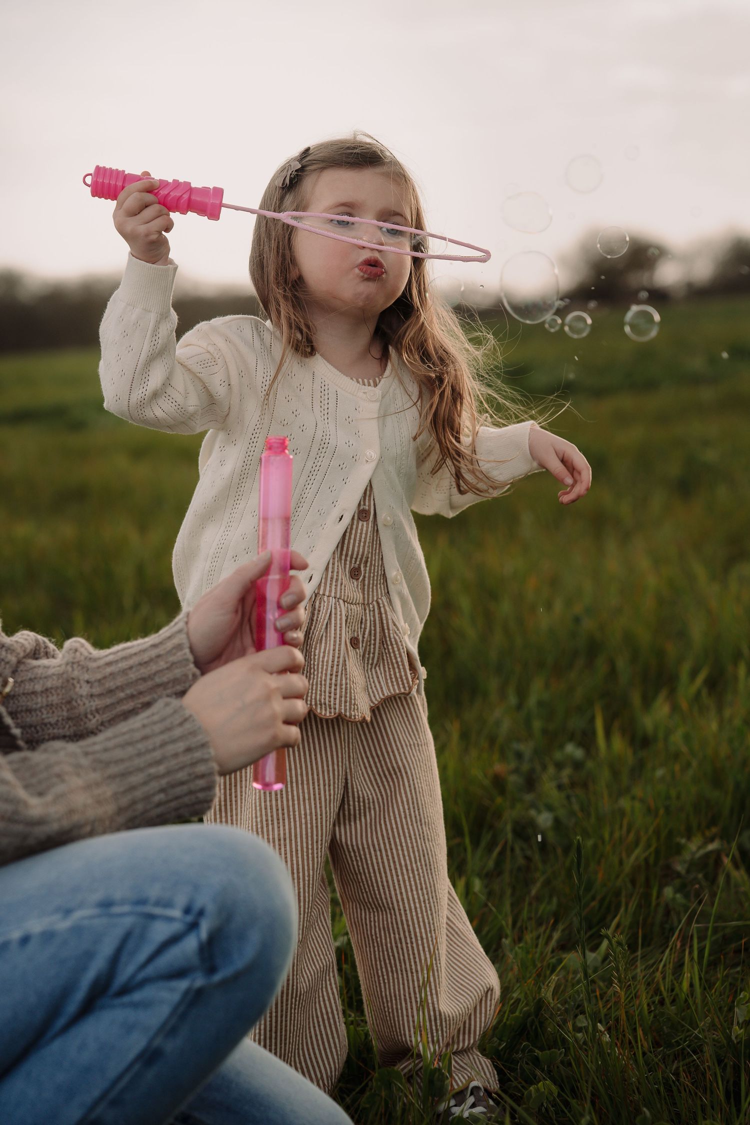 A young girl blowing soap bubbles outdoors with pink bubble wands on a green field.