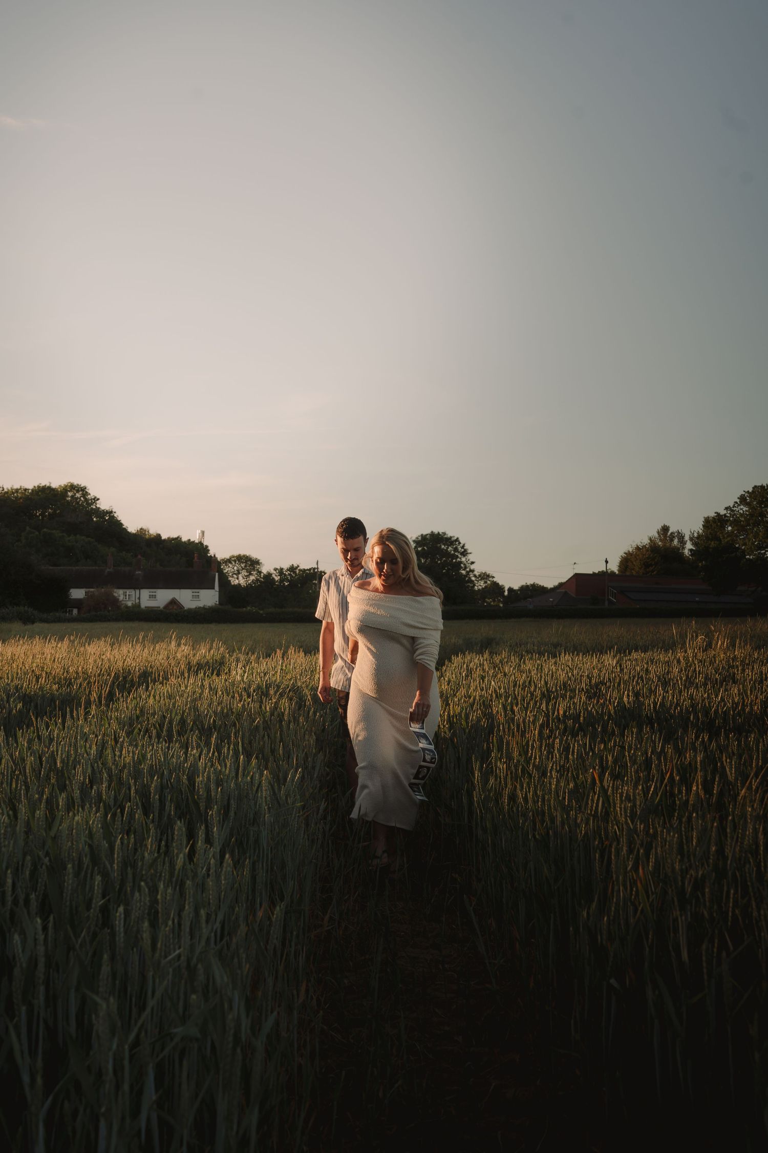 Couple embracing in a golden wheat field at sunset, with trees and a white farmhouse in the background.