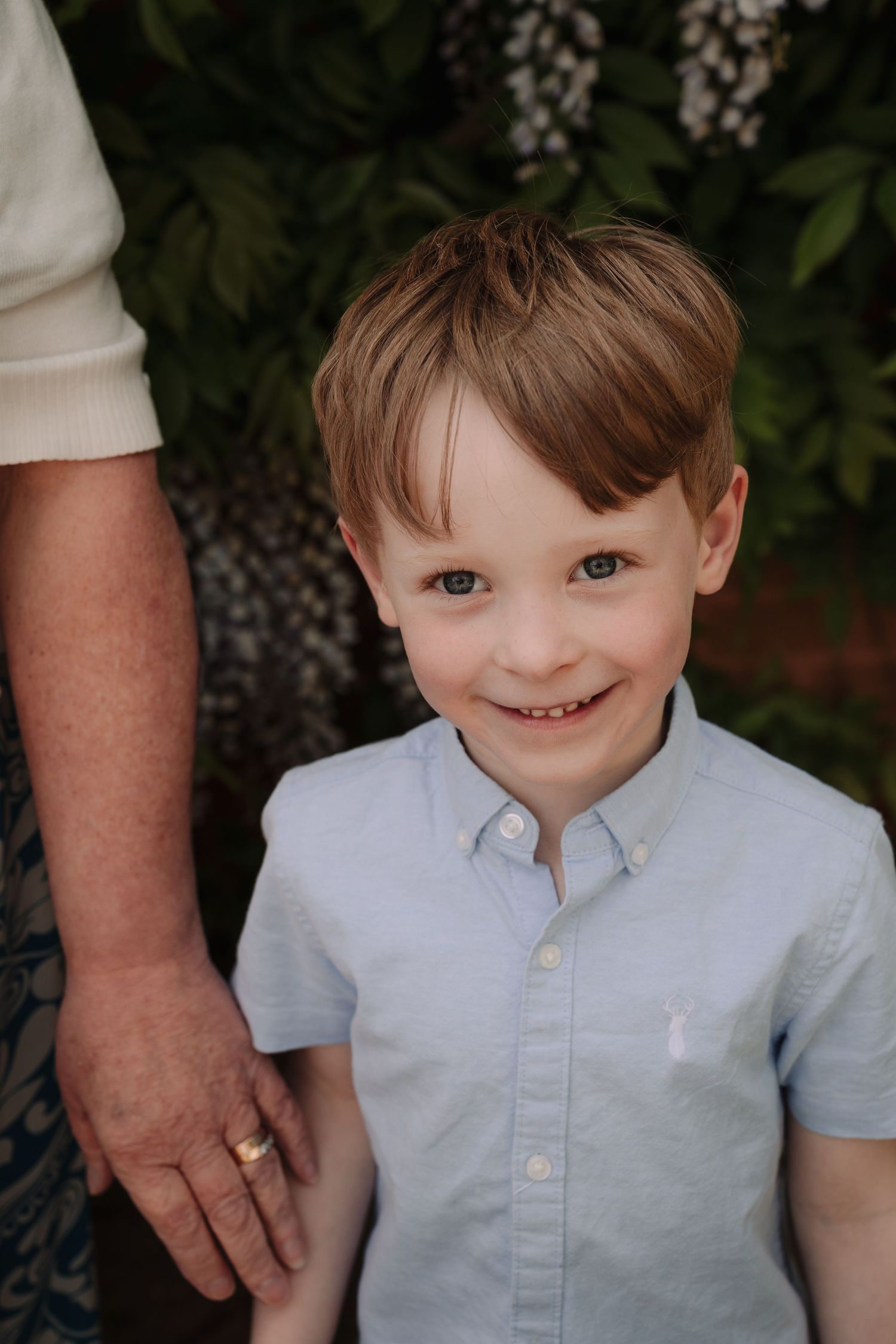 A smiling young boy with brown hair wearing a light blue button-up shirt holds an adult's hand outdoors.