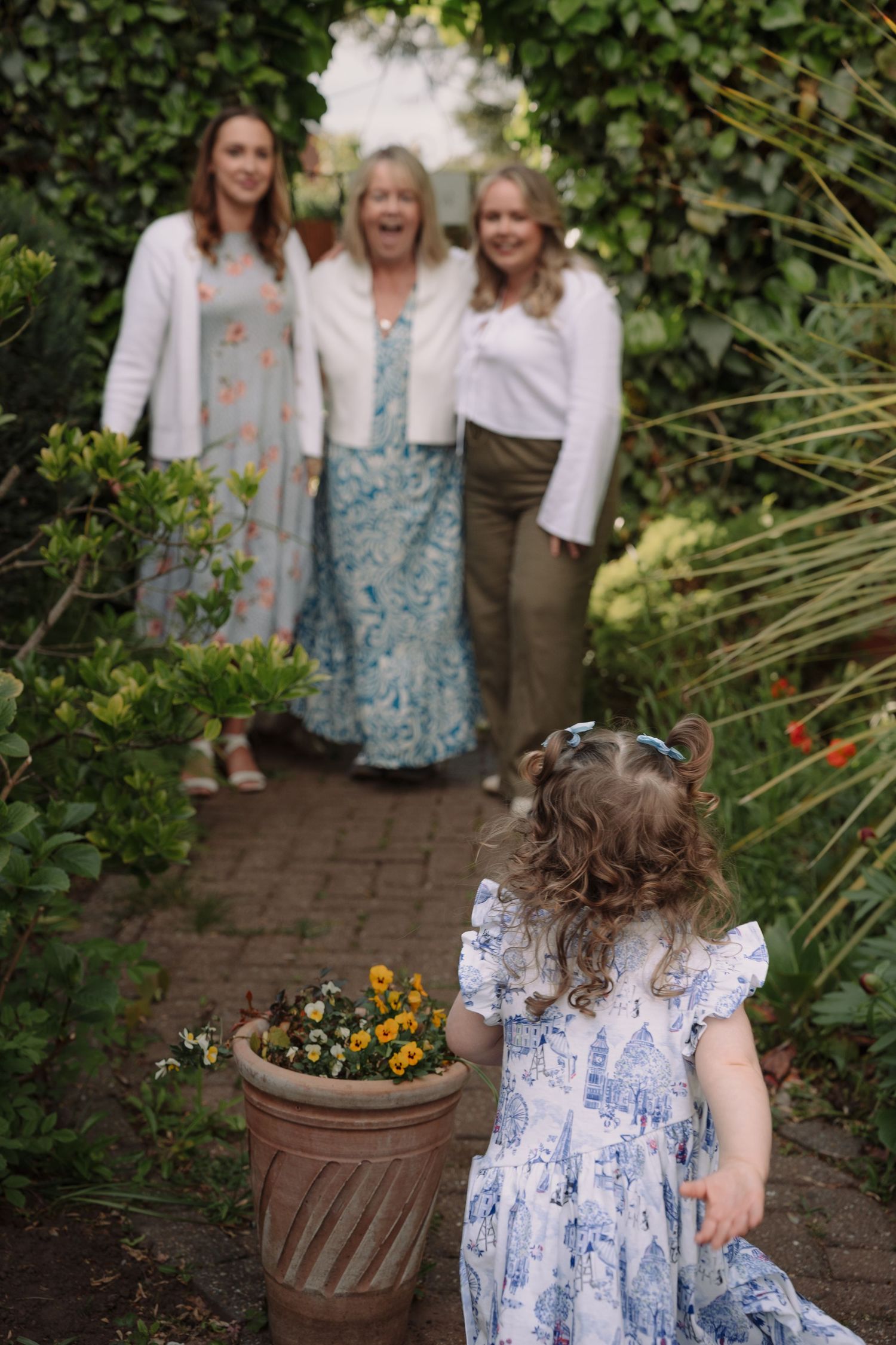 A little girl in a floral dress stands on a garden path facing three smiling women surrounded by lush greenery.