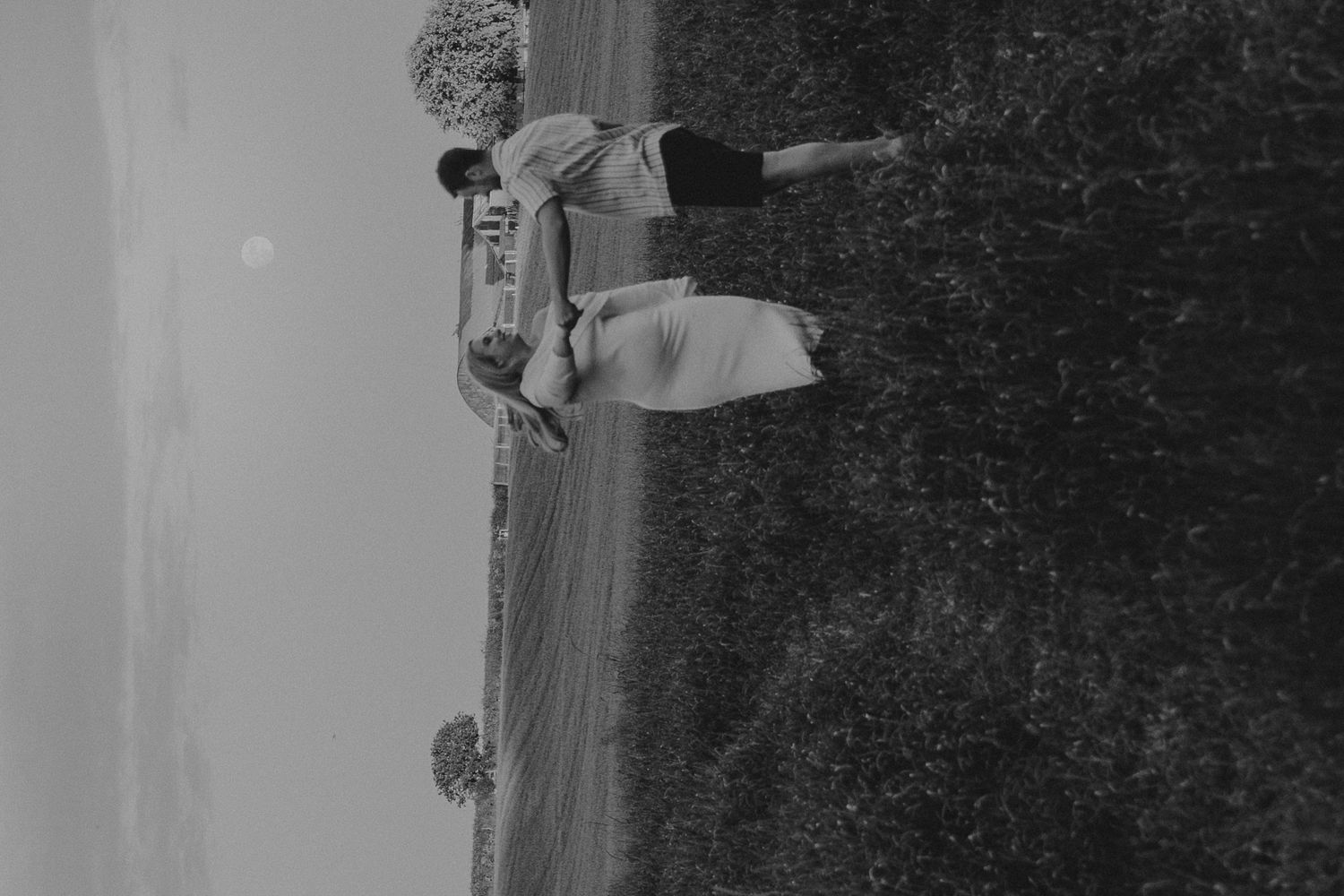 Black and white photo of two people embracing in an open field with a serene landscape in the background.