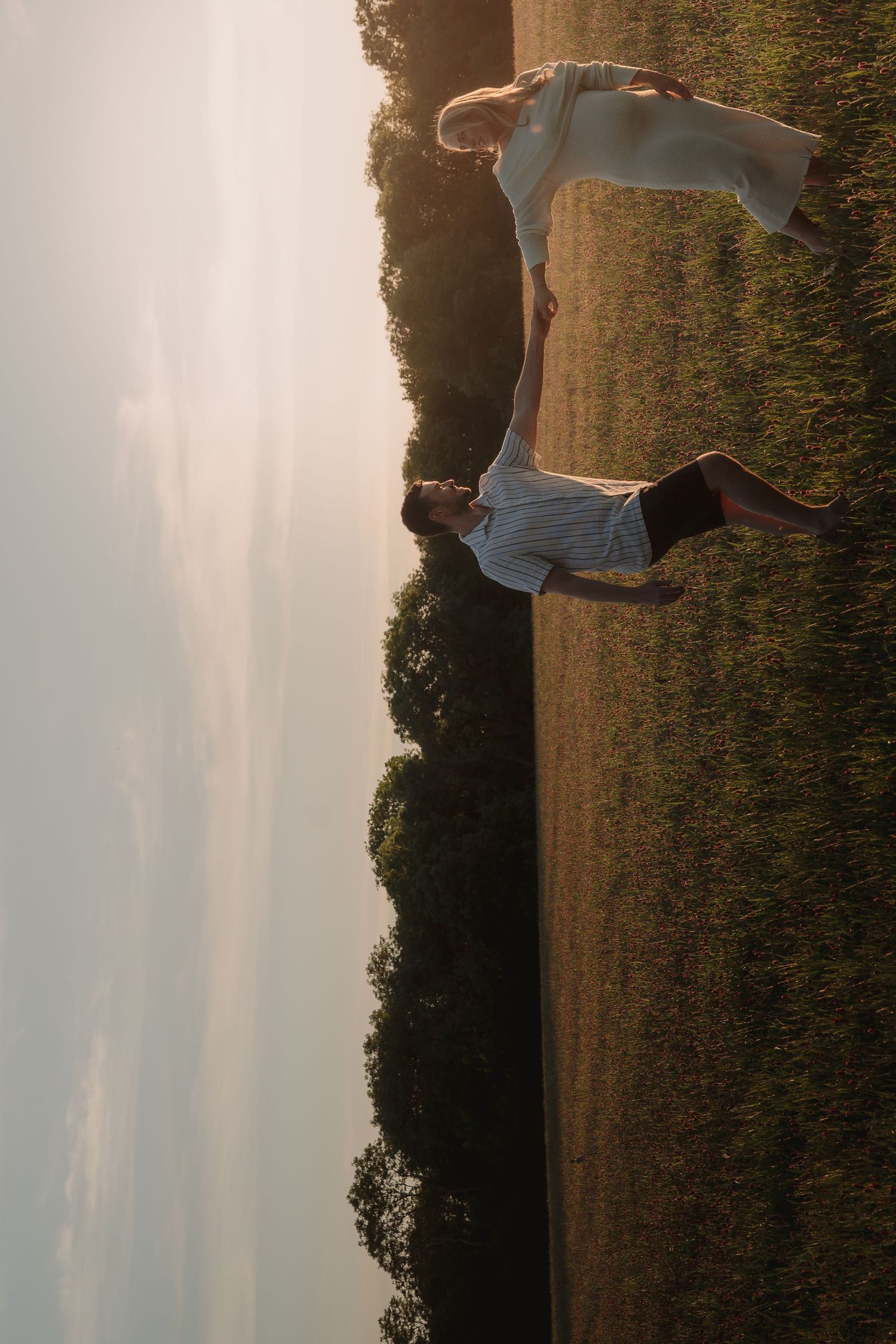 Couple dancing in a golden sunlit field at sunset, wearing white outfits, surrounded by lush green trees.