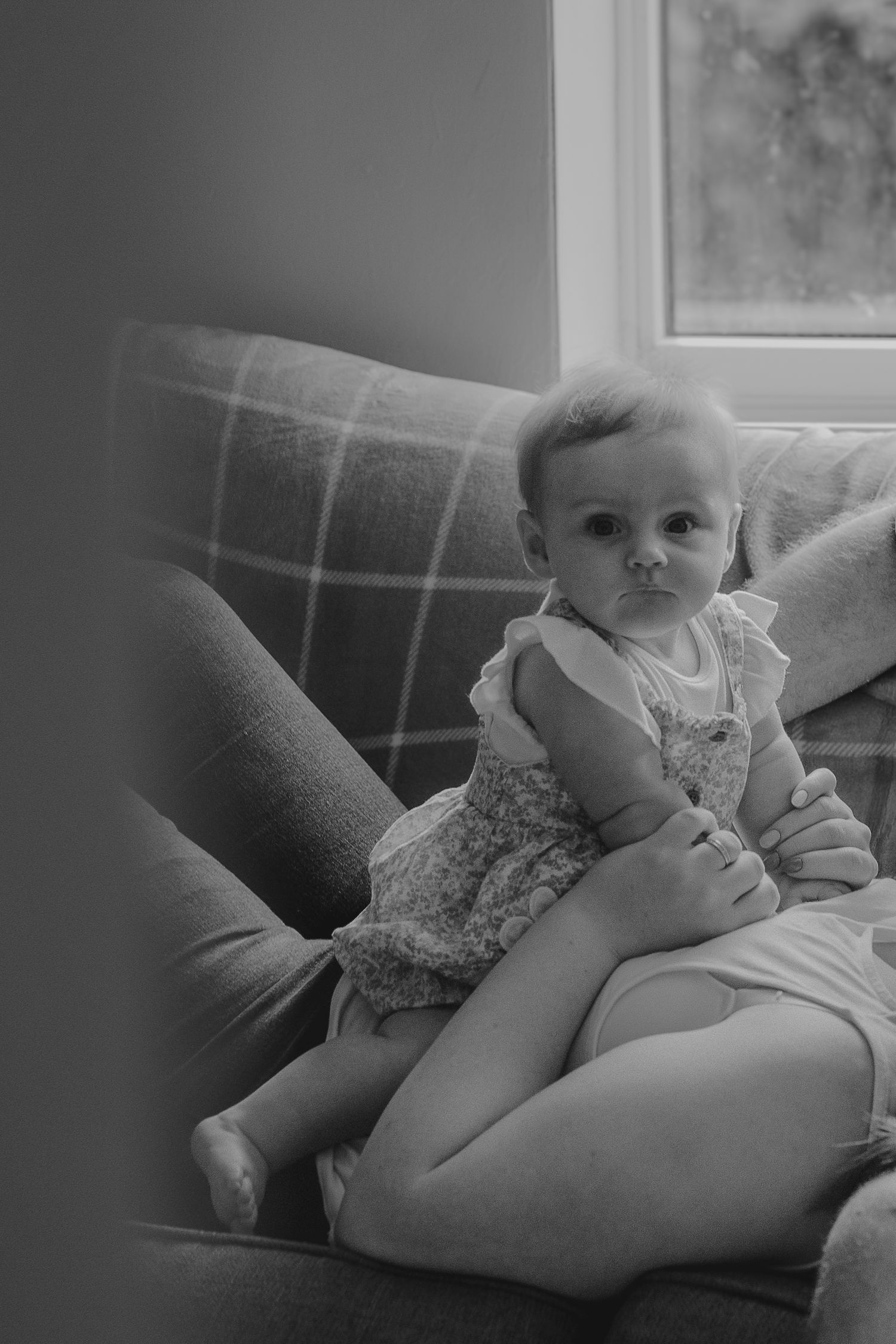 A black and white photo of a baby sitting on a plaid couch looking towards the camera.
