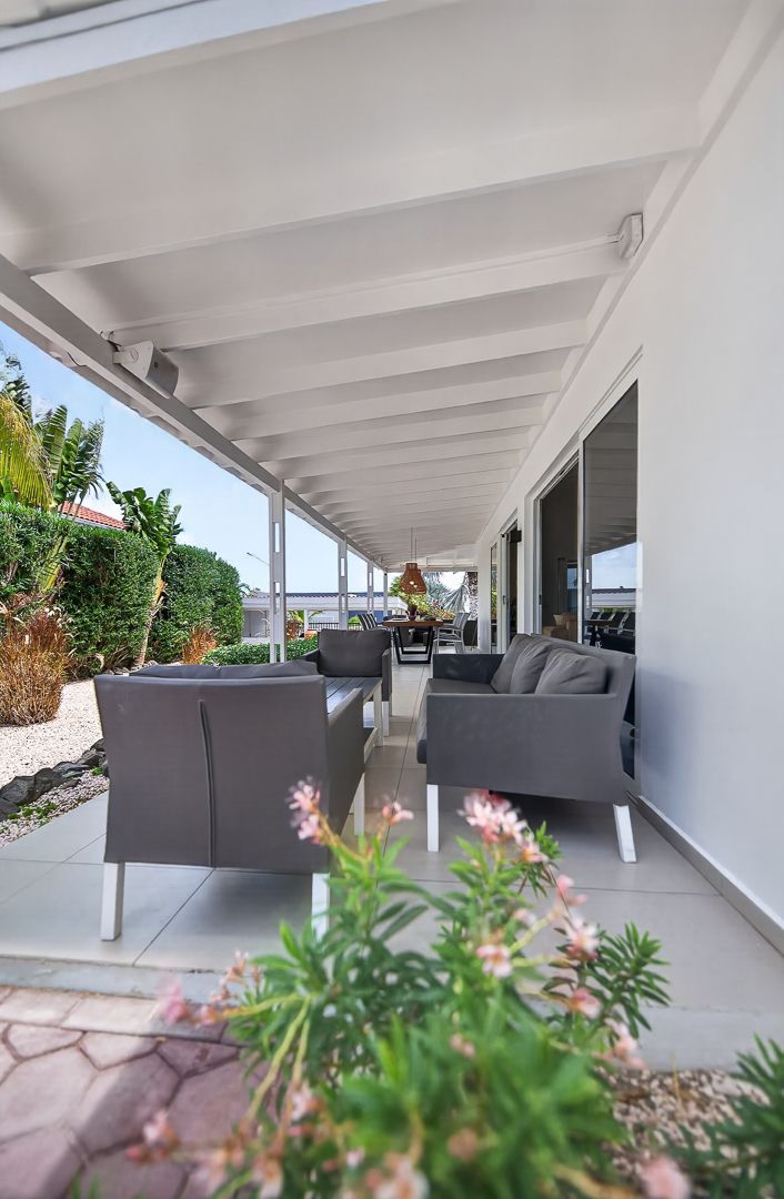 Covered outdoor patio with gray modern lounge chairs, white beams, and tropical garden views on a sunny day.