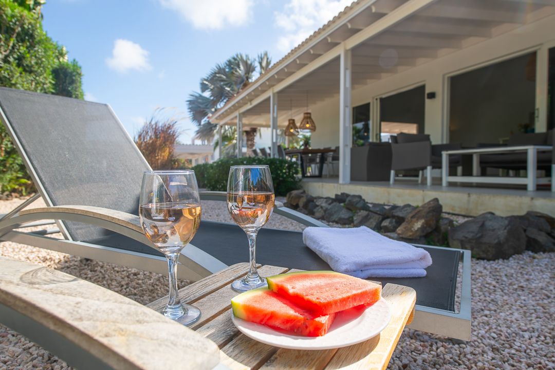 Two wine glasses with rosé and fresh watermelon slices on a plate beside poolside lounge chairs at a sunny villa.