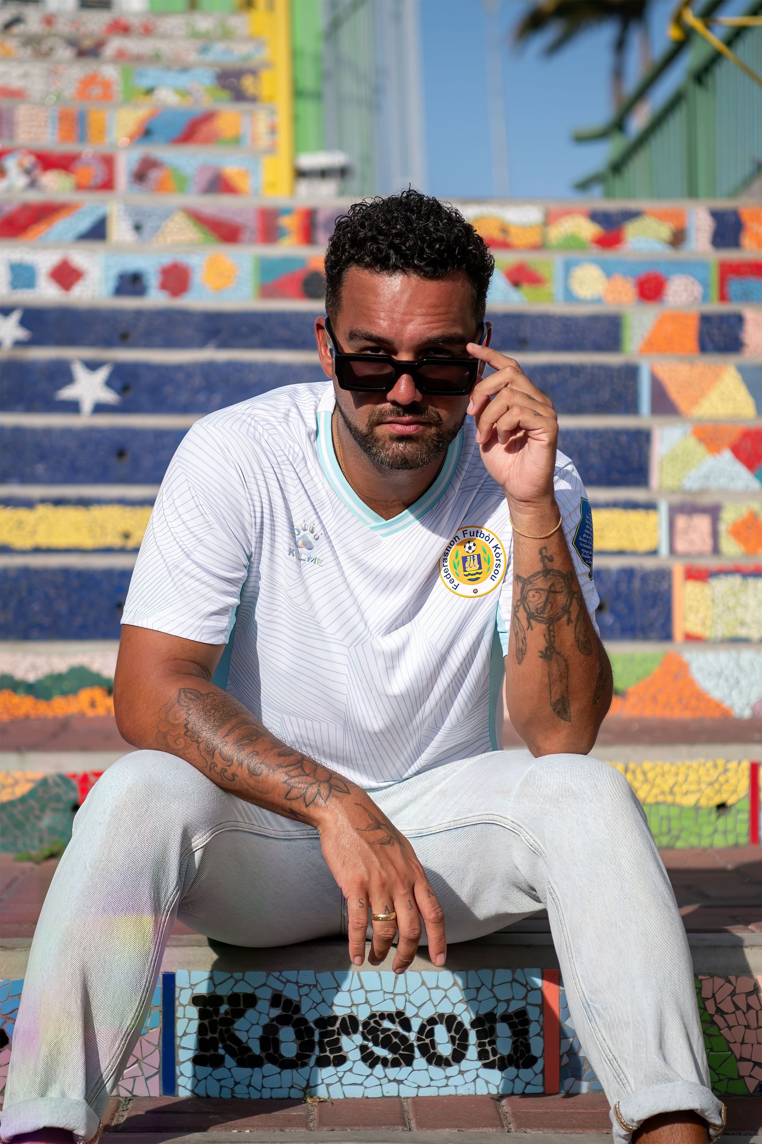 Man in white jersey sitting on colorful mosaic steps, wearing sunglasses, with tattoos visible.