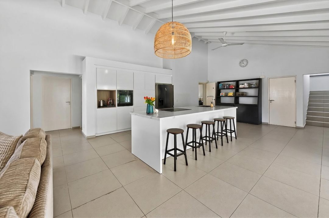 Modern open-plan kitchen with white island, bar stools, rattan pendant light, and large tile floors in a bright spacious interior.