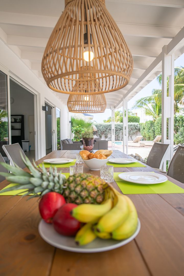 Tropical fruit platter with pineapple and bananas on outdoor dining table under rattan pendant lights at a villa.