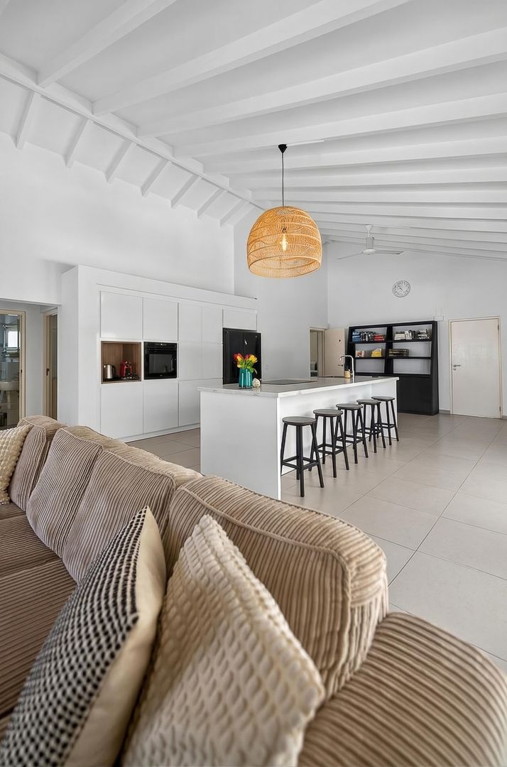 Modern open-plan living space with beige sofa, white kitchen island, bar stools, and rattan pendant light on vaulted ceiling.