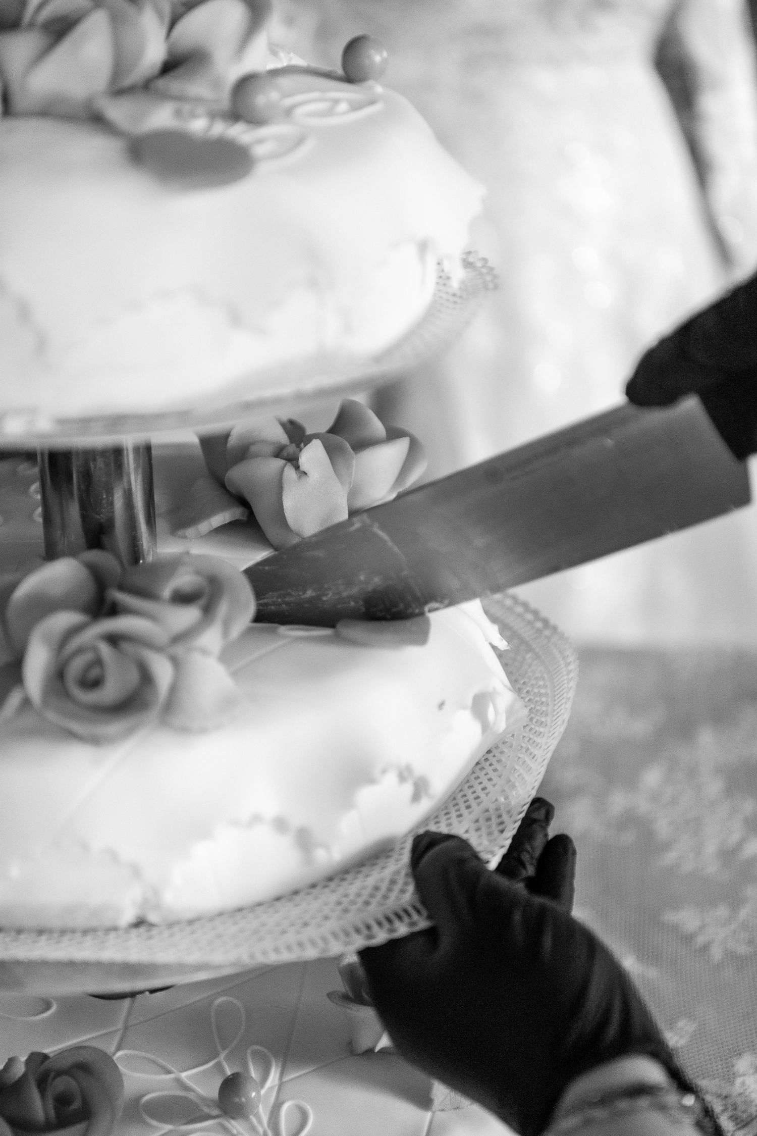 A large knife cuts into a white tiered wedding cake decorated with delicate roses and ribbons in black and white.