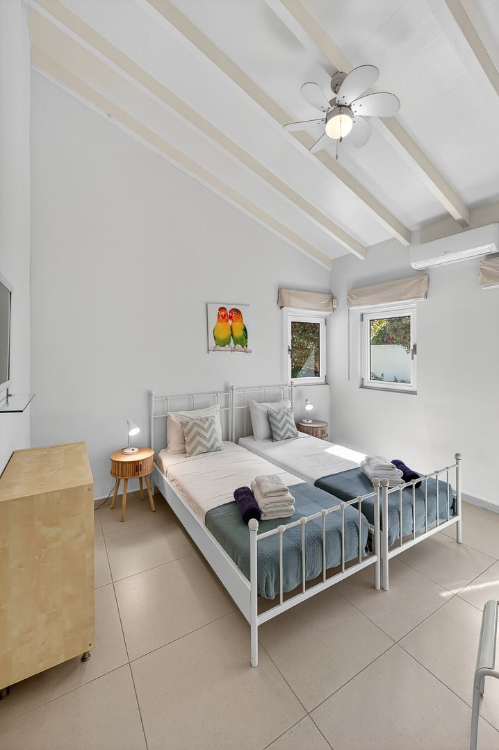 Bright white bedroom with twin beds, blue bedding, ceiling fan, exposed beams, and colorful parrot artwork on the wall.