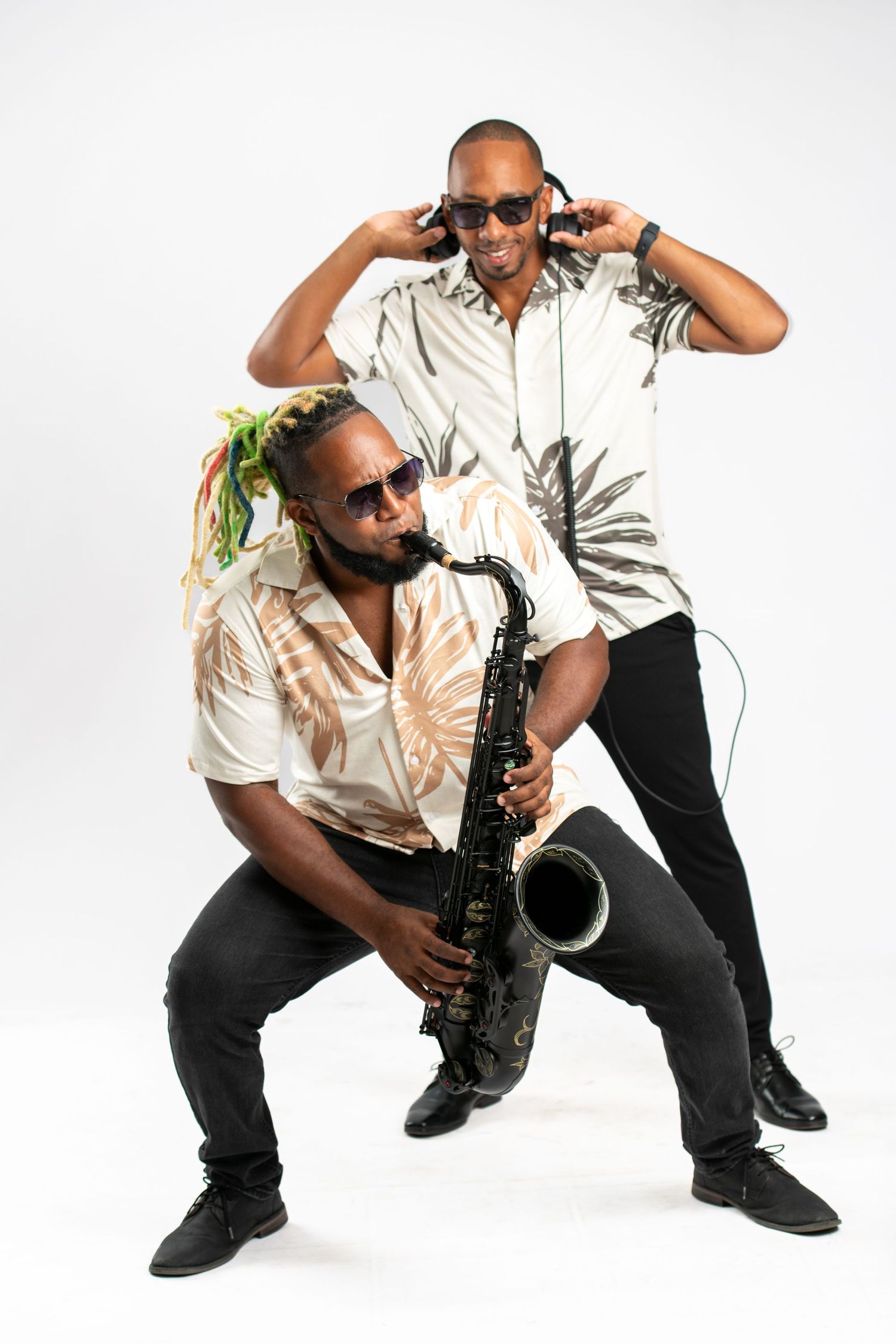 Two musicians pose together, one playing saxophone and one wearing DJ headphones, in tropical shirts on white background.