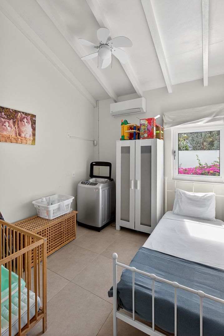 Bright bedroom with white metal bed, ceiling fan, wardrobe, washing machine, baby crib, and AC unit near a sunny window.