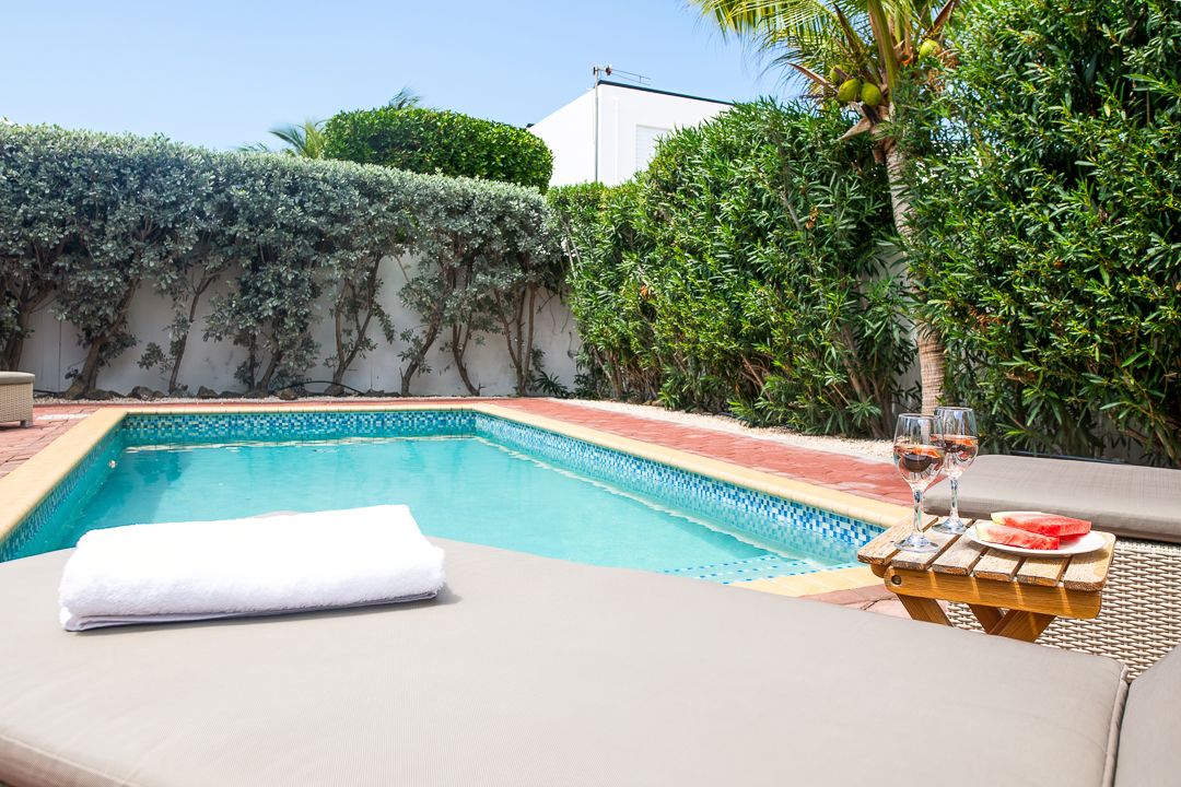 Luxury outdoor pool with lounge chair, wine glasses, and lush green hedges on a sunny day.