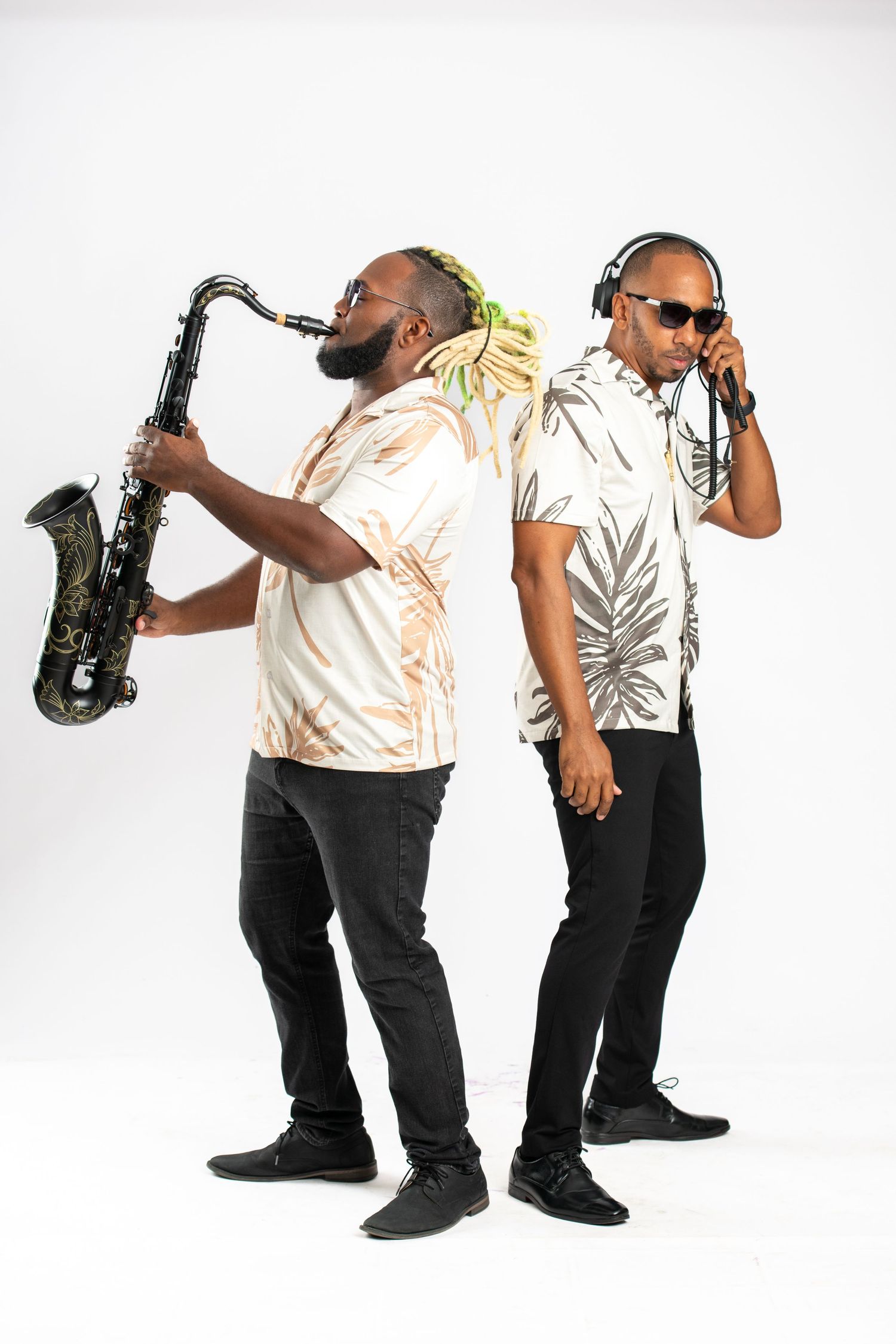 Two male musicians pose back-to-back, one playing saxophone, one wearing headphones, in tropical shirts on white background.