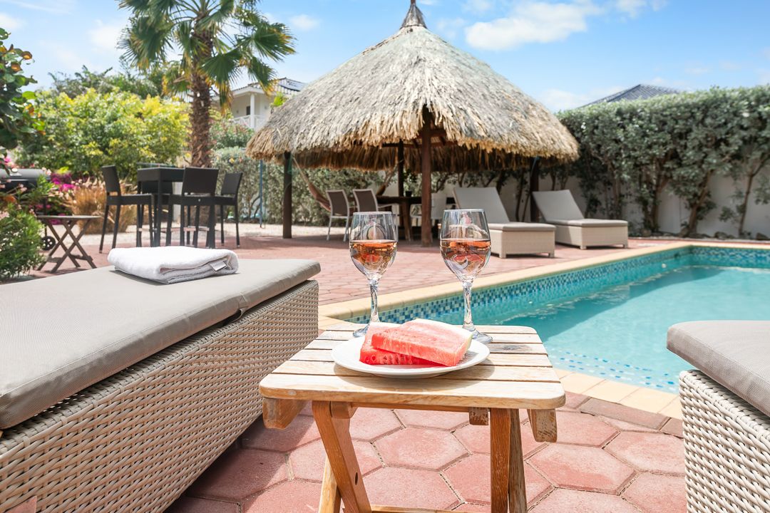 Tropical poolside setting with wicker loungers, thatched palapa, rose wine, and fresh watermelon slices.