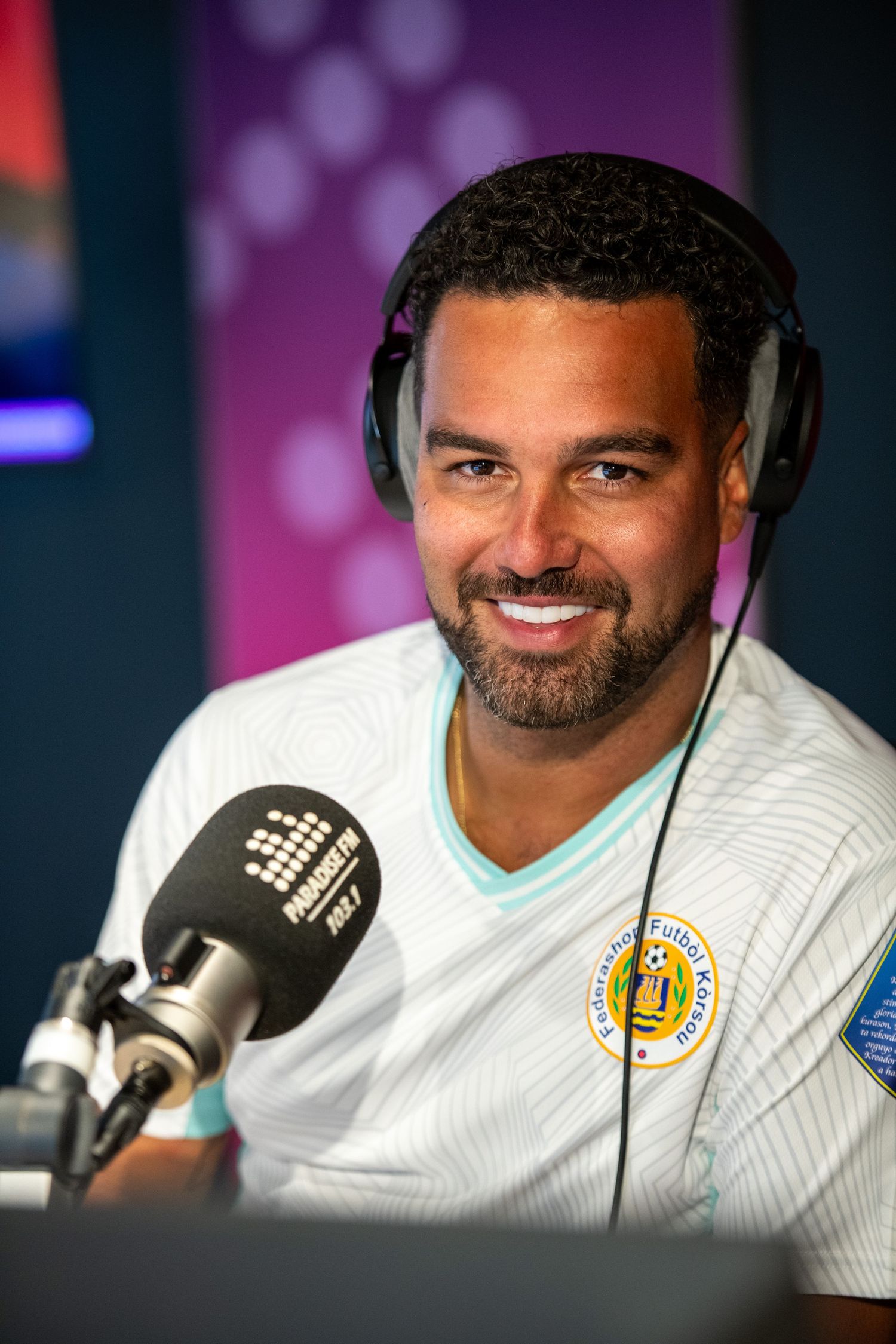 Smiling man wearing headphones and a white soccer jersey speaks into a microphone at a radio studio.