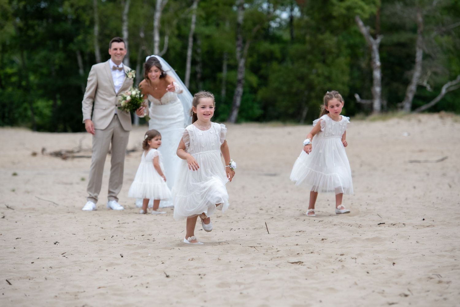 A wedding party walks down a sandy beach with flower girls in white dresses and a bride carrying a bouquet.