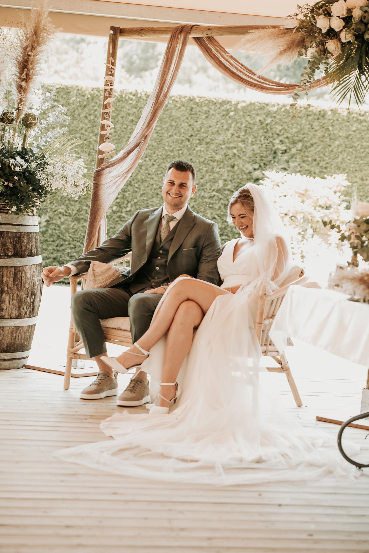 A couple sits together on a rustic porch decorated with flowy fabric and greenery for their wedding celebration.