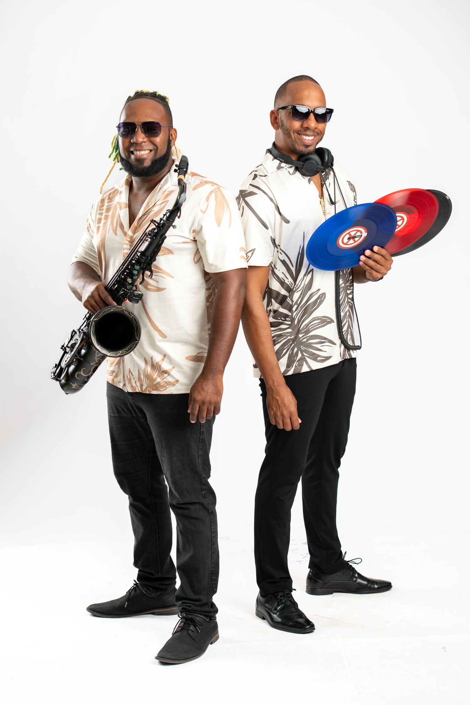 Two smiling musicians in tropical shirts, one holding a saxophone and one holding colorful vinyl records with headphones.