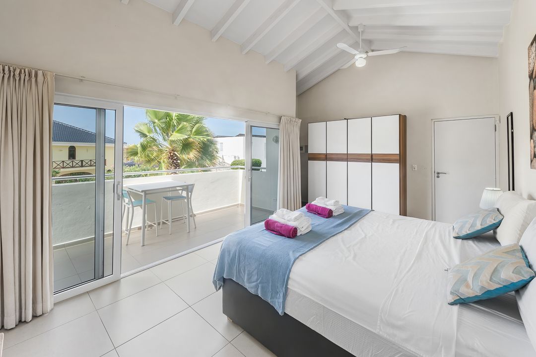 Bright bedroom with king bed, colorful pillows, wardrobe, and balcony with palm tree view.