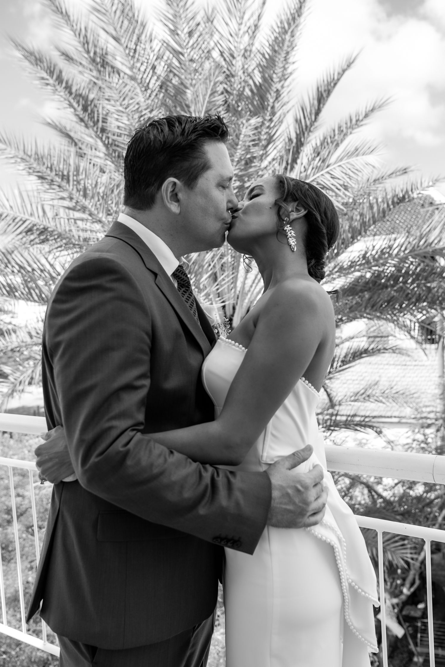 A romantic wedding couple shares a tender kiss under palm trees during their outdoor tropical ceremony.