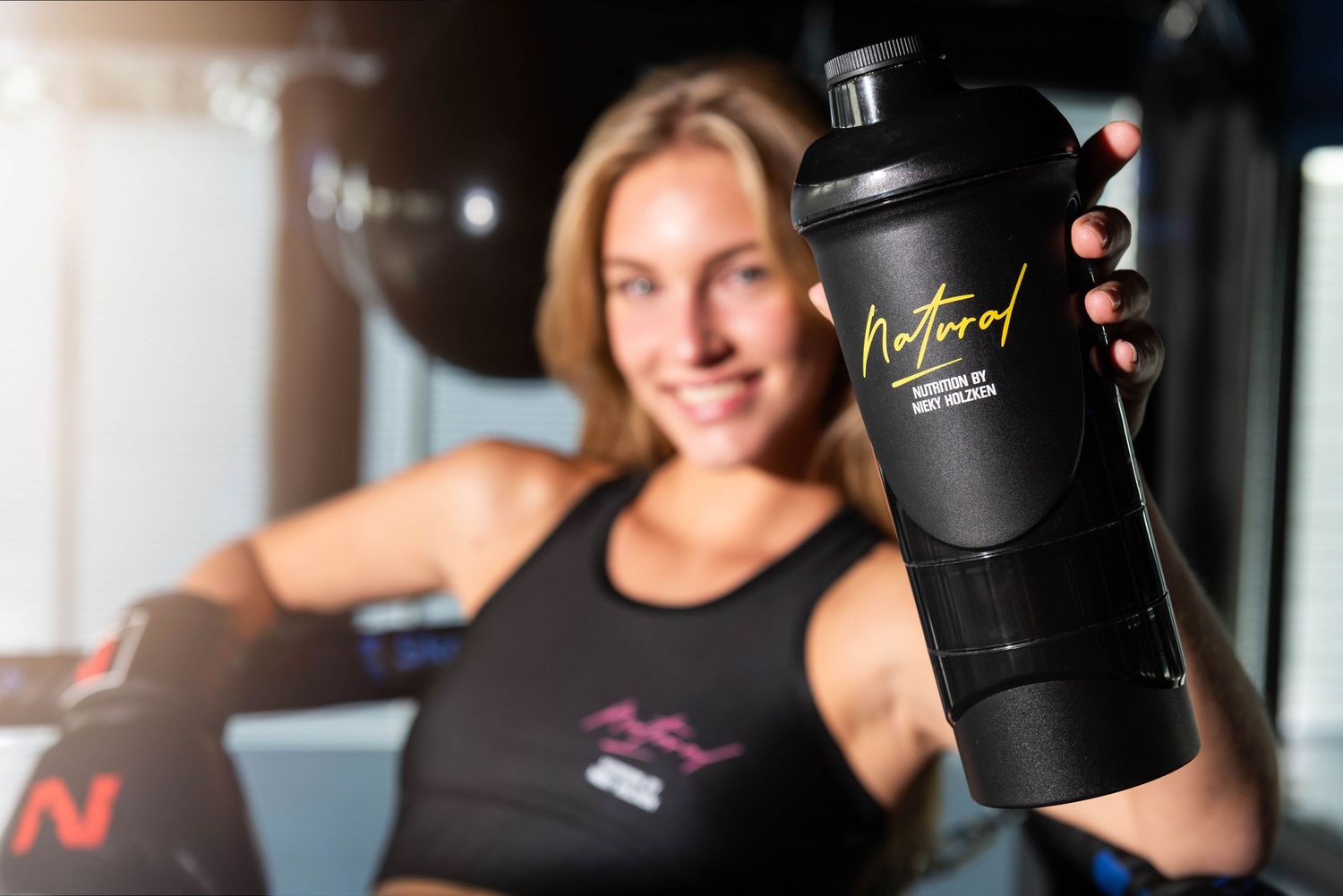A row of black shaker bottles and protein supplements lined up on a counter with gym equipment in the background.